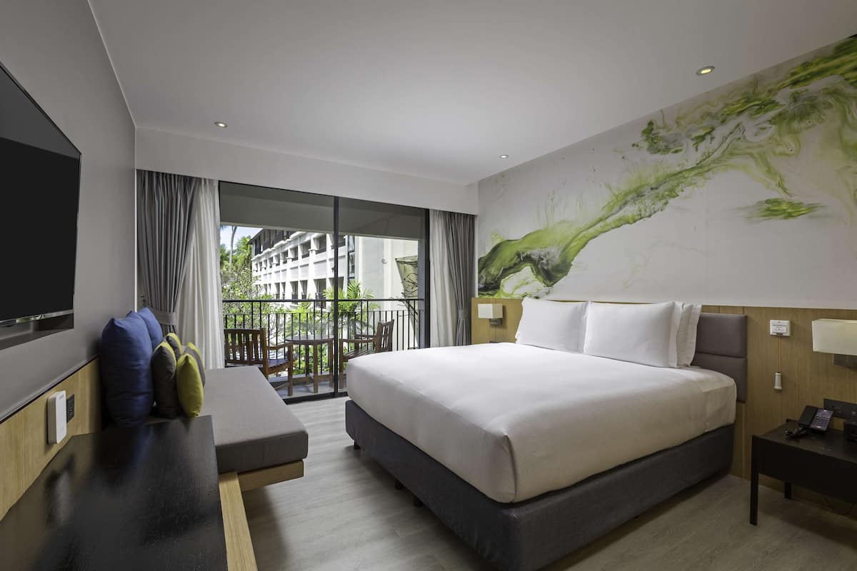 Habitación de hotel moderna con una cama grande, un televisor de pantalla plana, un sofá y un balcón con vistas a edificios y zonas verdes.