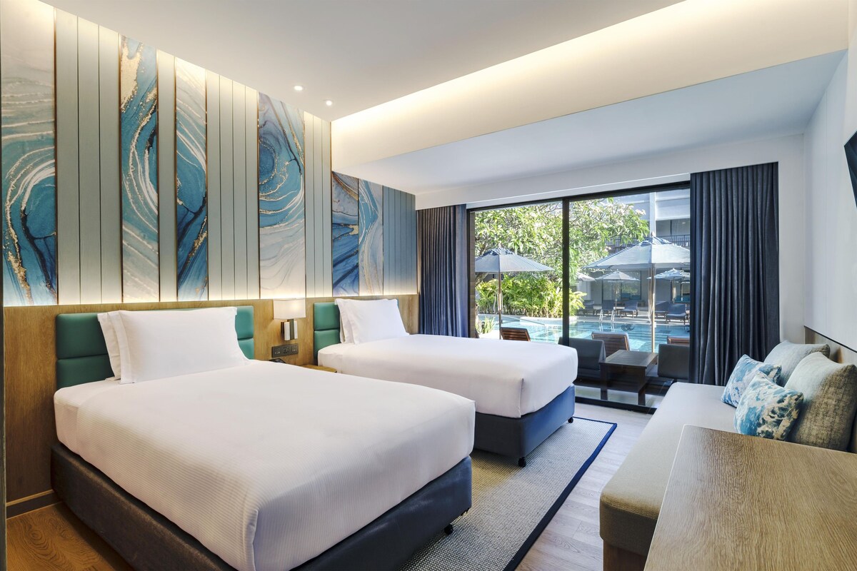 A hotel room with two beds, a sofa, a large window with a view of a pool and outdoor seating, and a wall with abstract art.