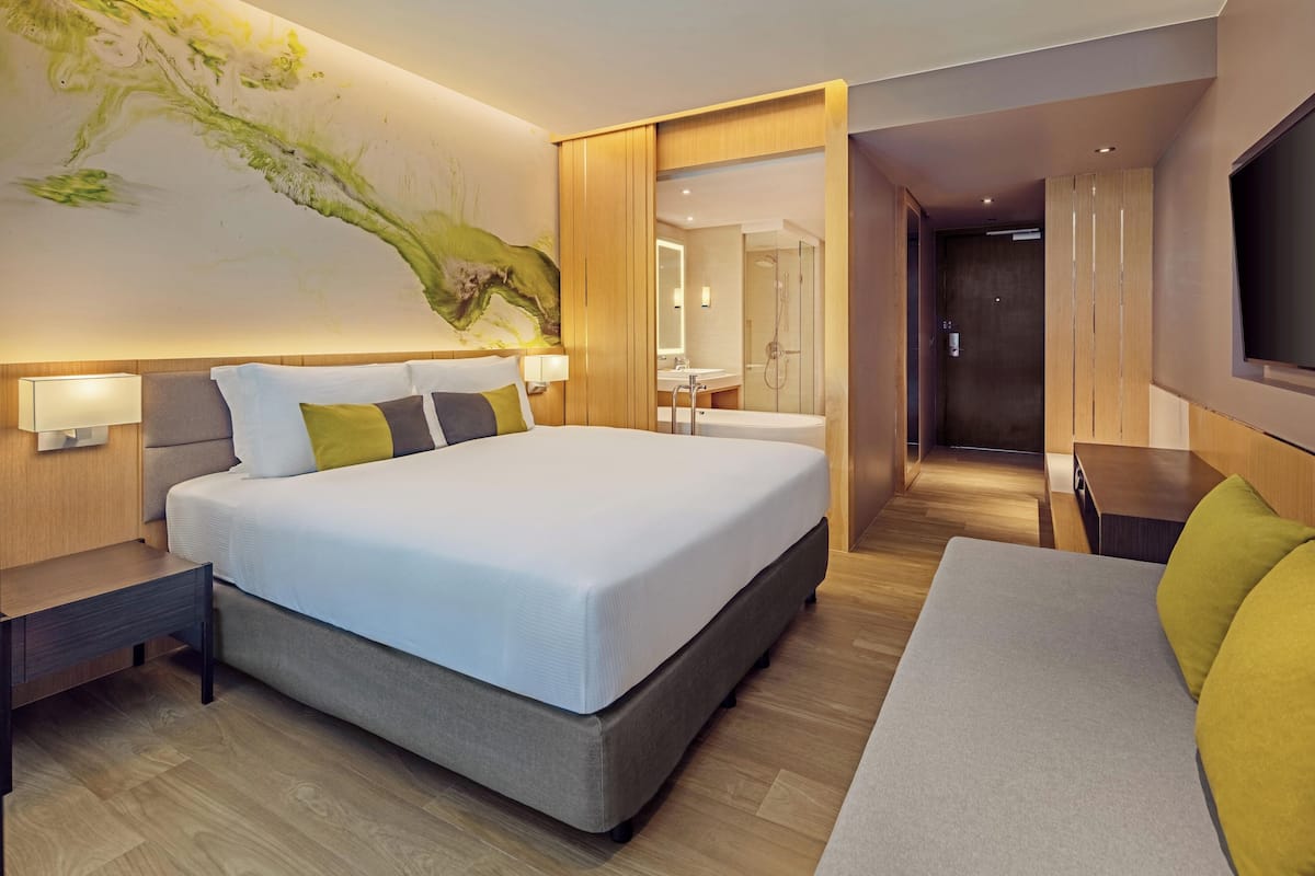 Una habitación de hotel moderna con una cama grande, mesitas de noche, un sofá, un baño con ducha y un televisor montado en la pared.