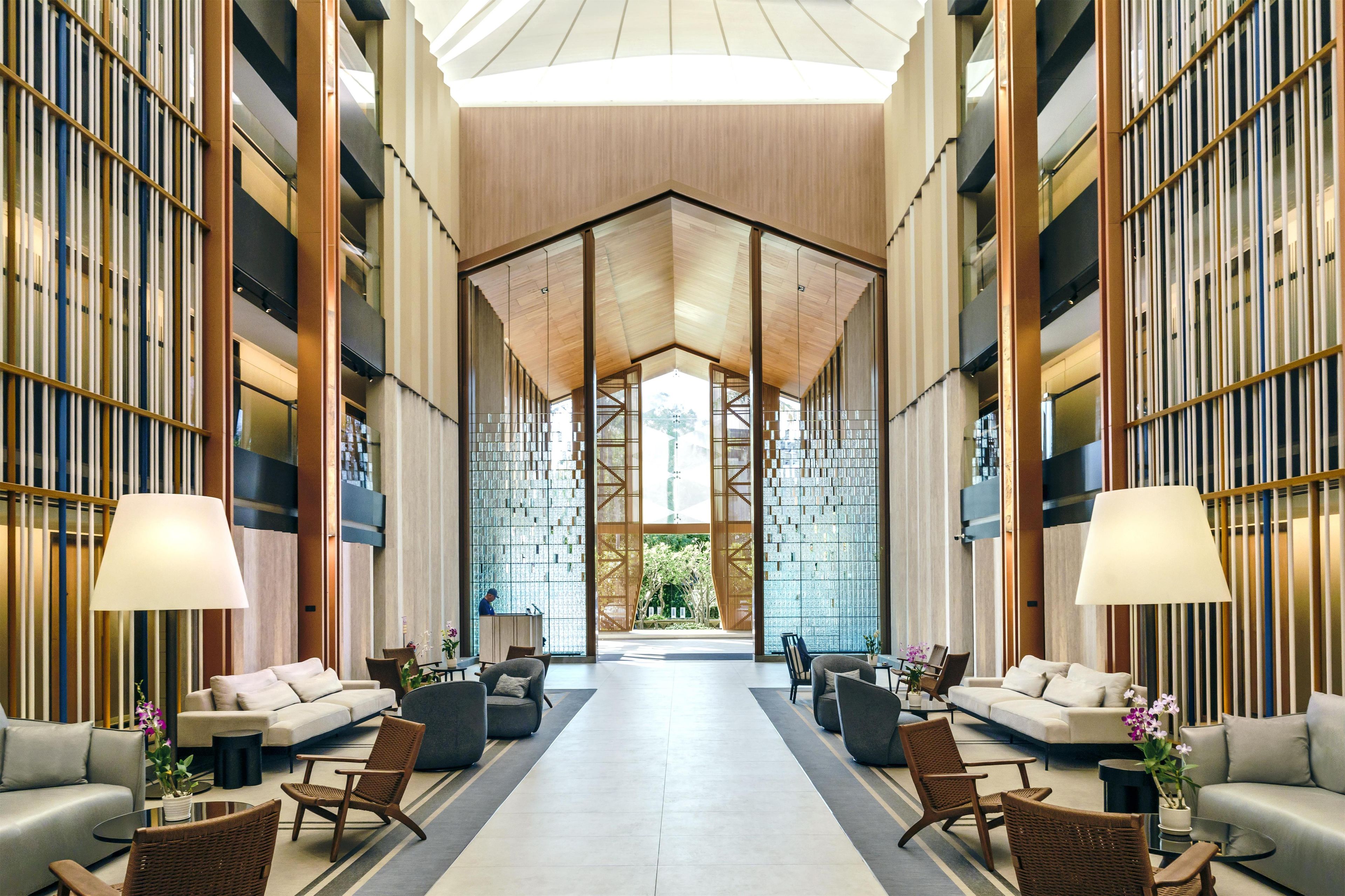 A spacious lobby with a high ceiling, a central seating area with sofas and chairs, and a large glass wall providing a view of the outdoors.