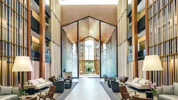 A spacious lobby with a high ceiling, a central seating area with sofas and chairs, and a large glass wall providing a view of the outdoors.