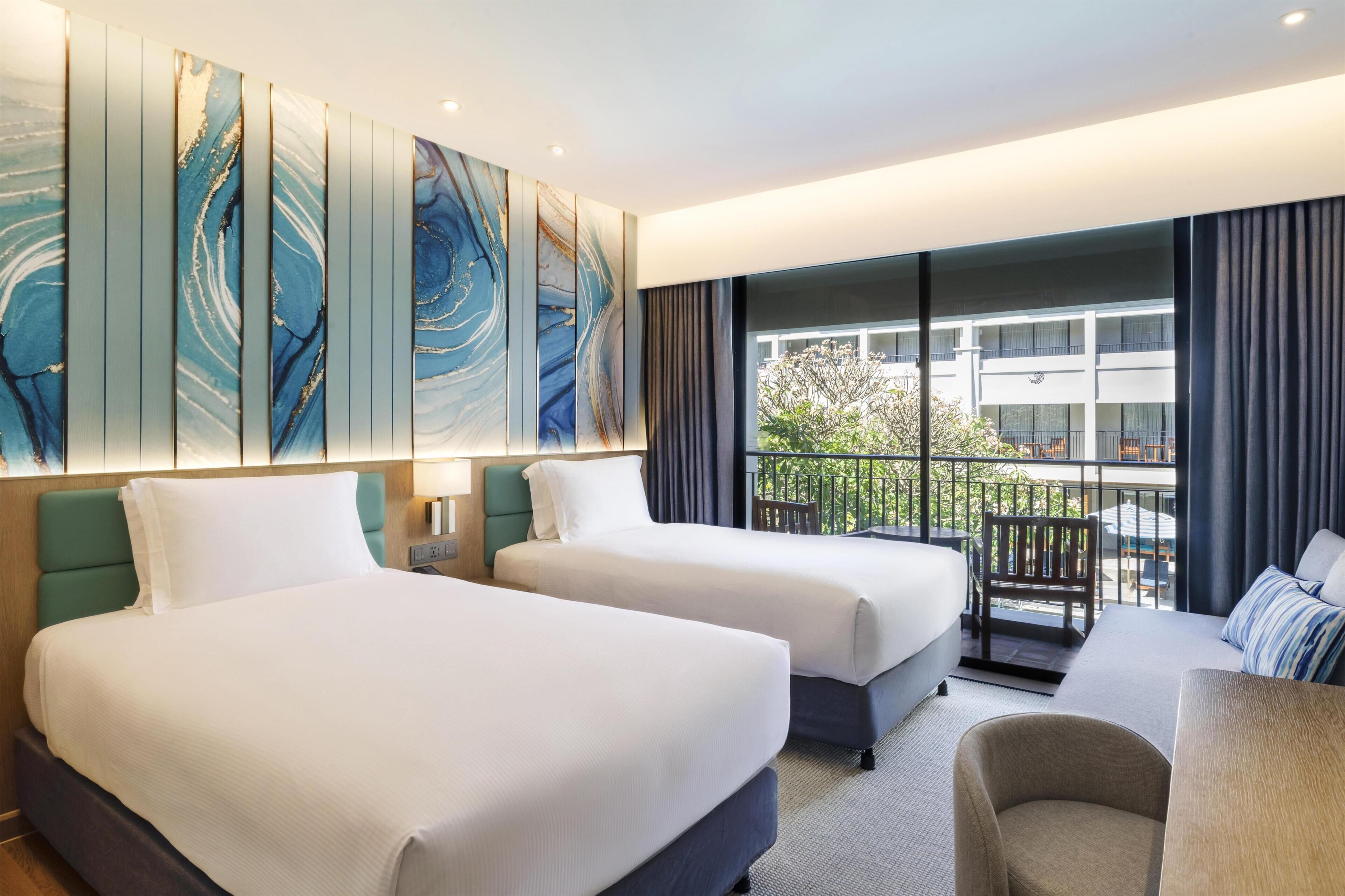 A hotel room with two beds, a sofa, a balcony with a view of a building, and a large abstract painting on the wall.
