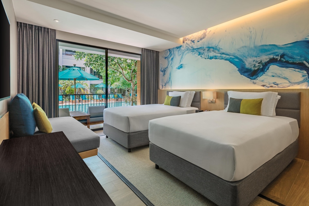 A hotel room with two beds, a desk, and a view of an outdoor area with umbrellas.