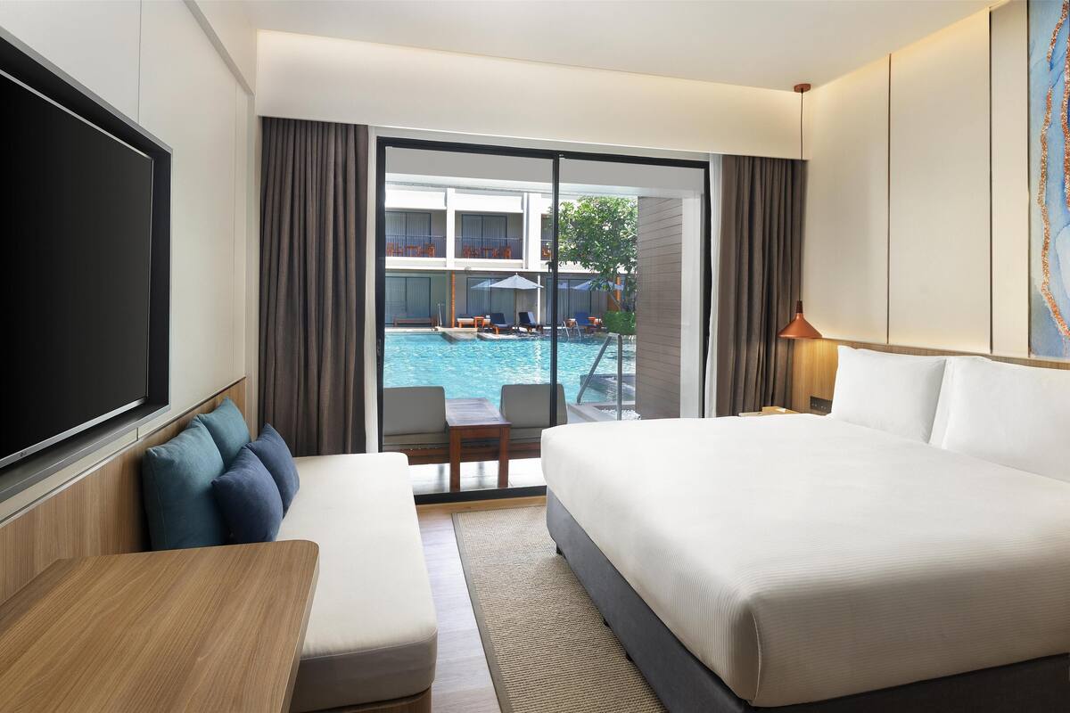 Habitación de hotel con una cama grande, un sofá, un televisor y vistas a una piscina y un edificio.
