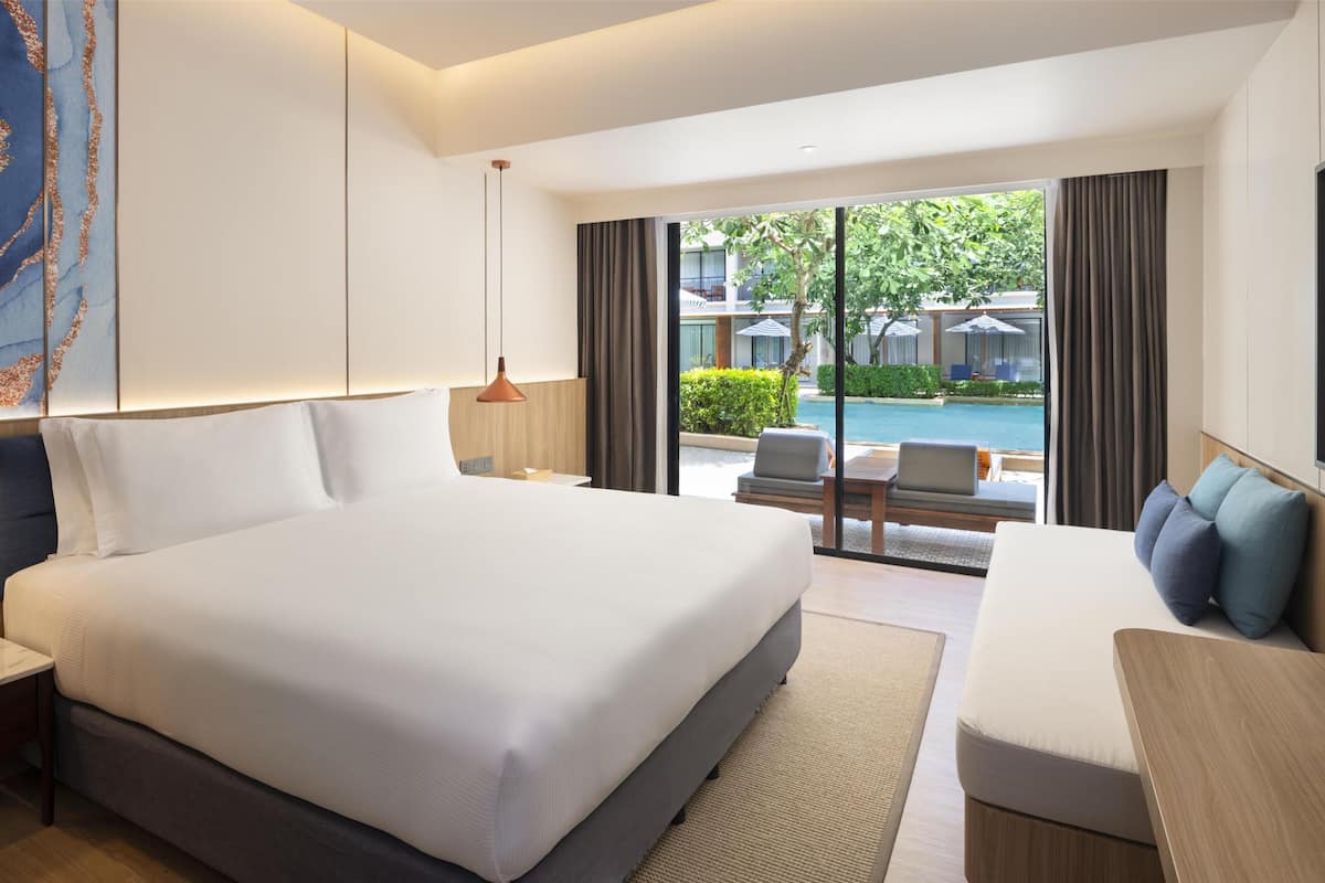 Habitación de hotel moderna con una cama grande, un sofá y vistas a una piscina y zona de estar exterior.