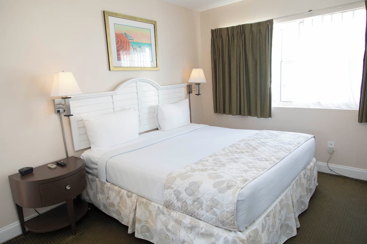 A neatly made bed with a white headboard and a floral-patterned bed skirt. There is a nightstand with a lamp and a remote control beside the bed. A painting hangs on the wall above the bed.