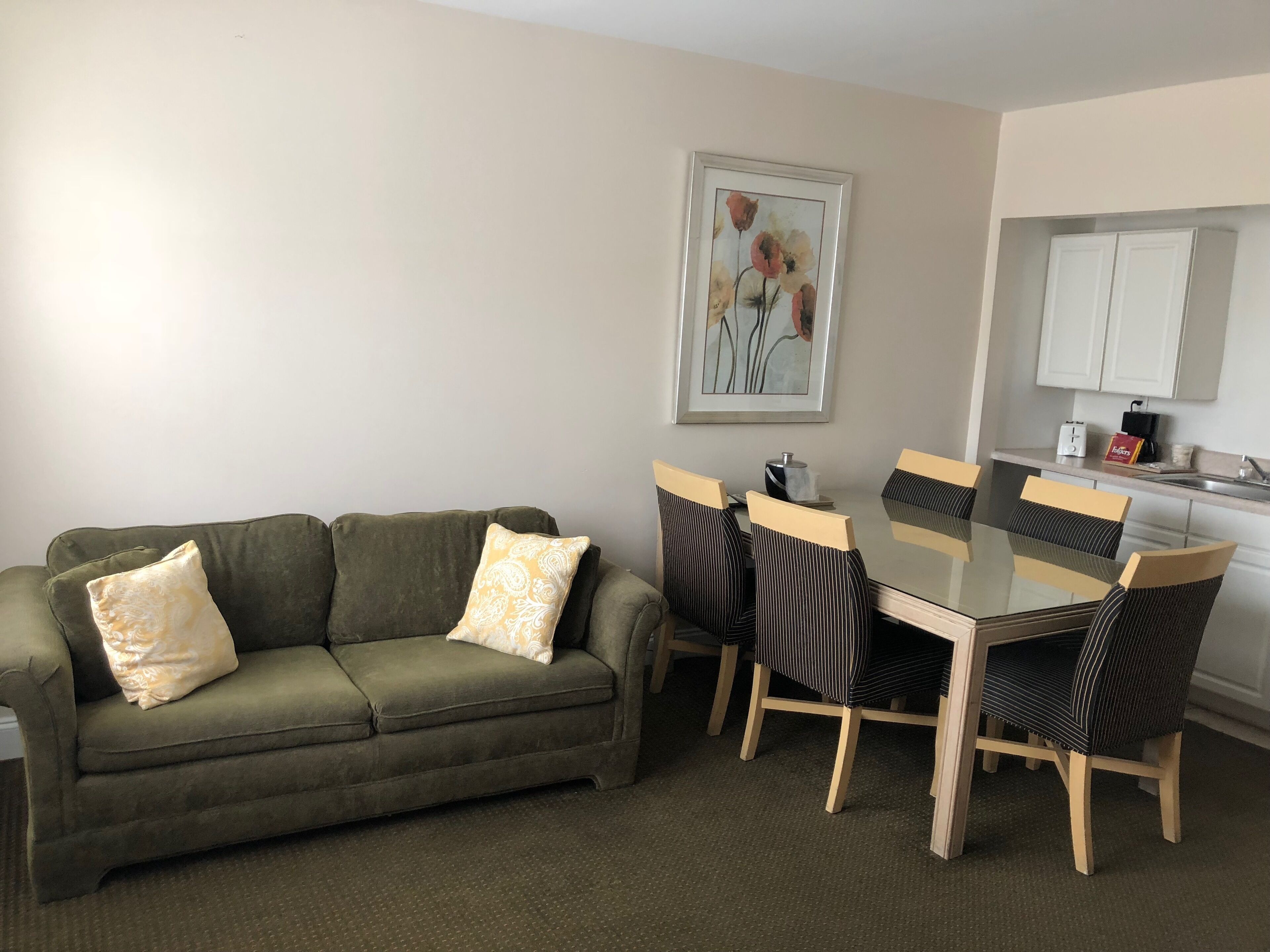 A living room with a green sofa, a glass-top dining table, and striped chairs. There is a framed floral artwork on the wall, and a kitchen area with white cabinets and a sink.