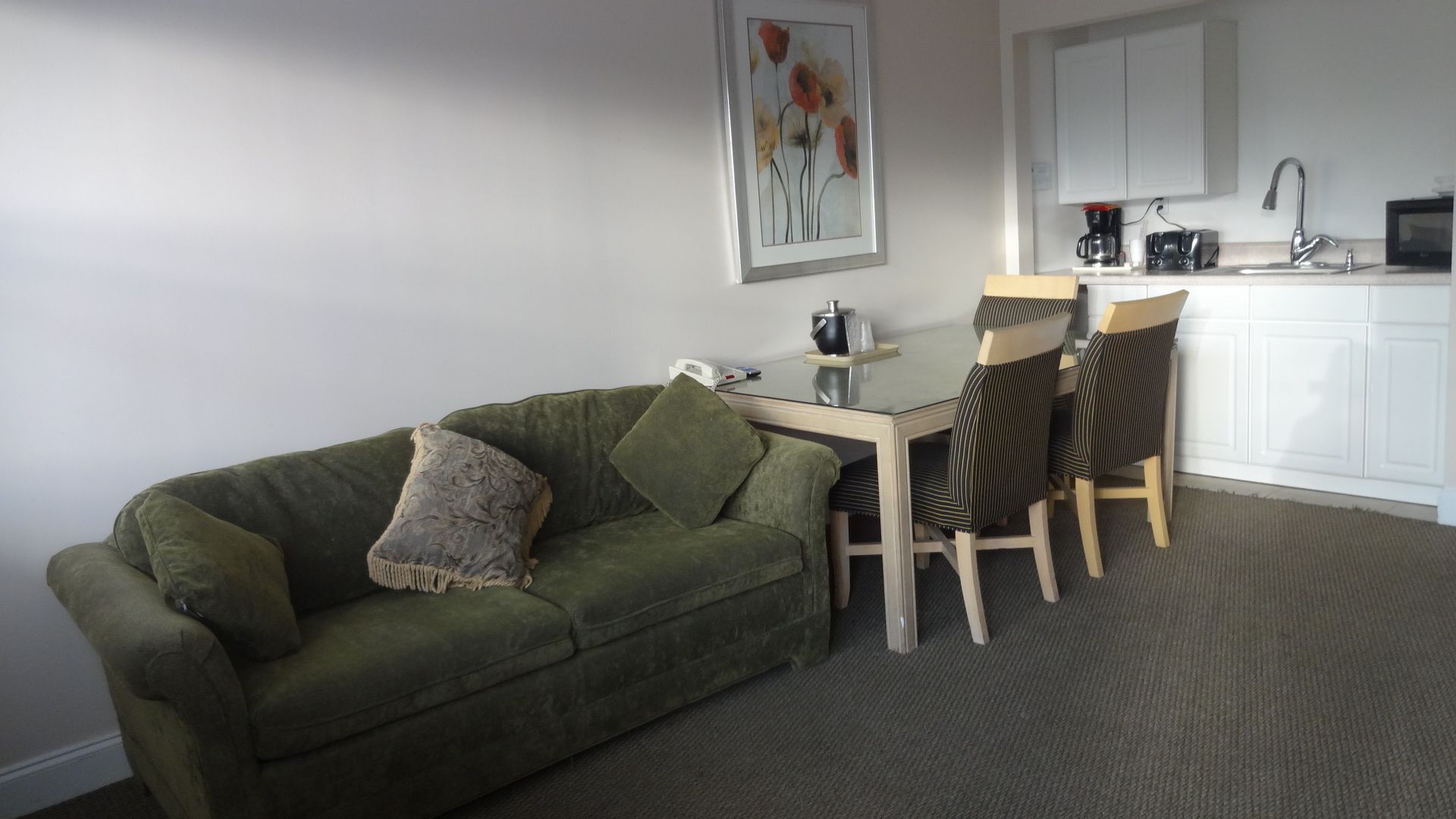A small living area with a green sofa, a coffee table, and two striped chairs. There is a kitchen area with white cabinets and a microwave.