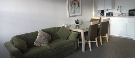A small living area with a green sofa, a coffee table, and two striped chairs. There is a kitchen area with white cabinets and a microwave.