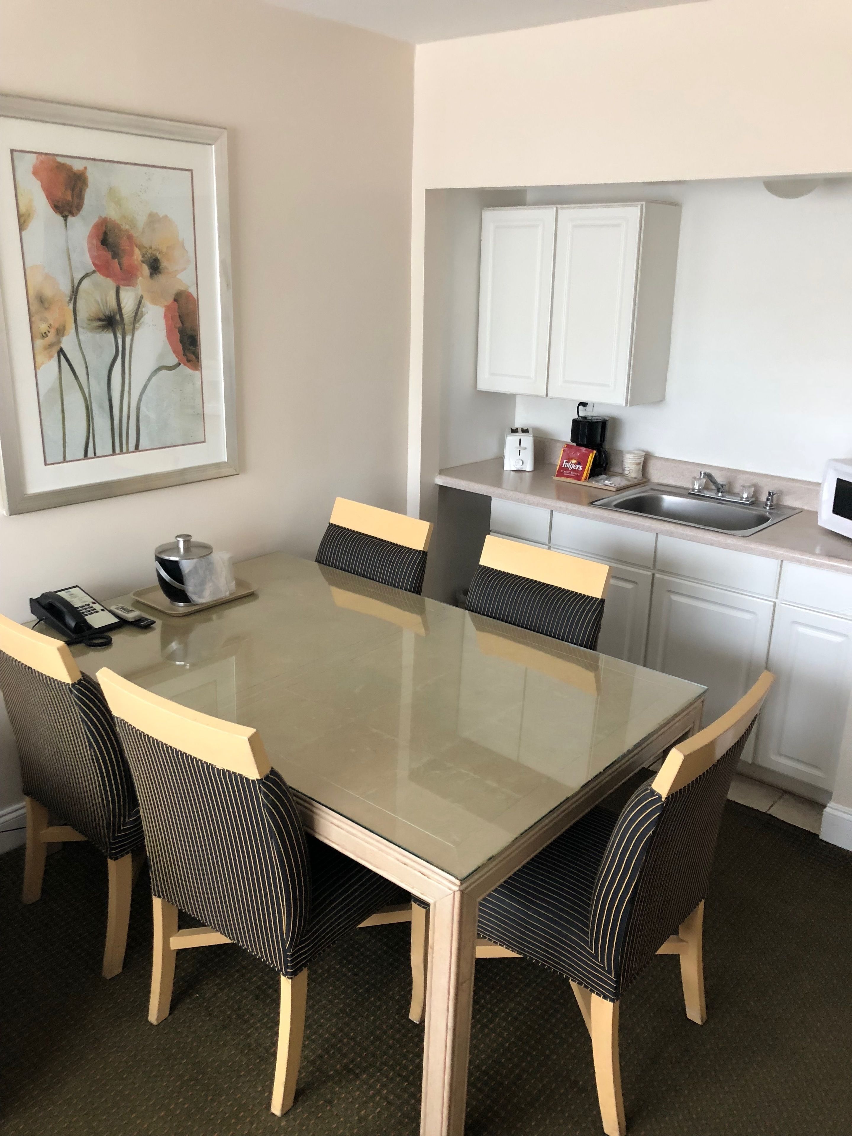 A dining area with a glass table and striped chairs, a kitchen with white cabinets and a sink, and a framed floral artwork on the wall.