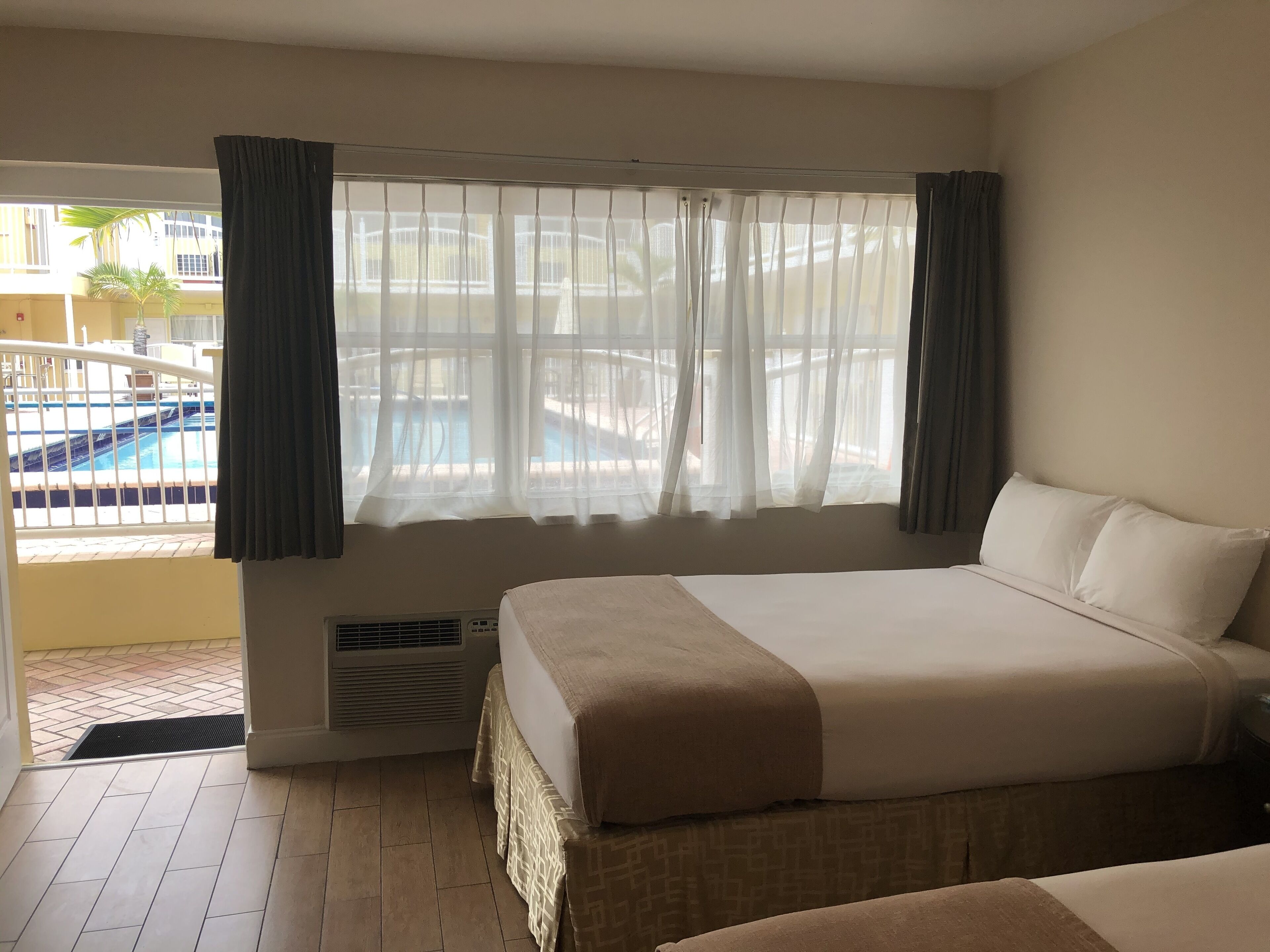 Een hotelkamer met een groot bed, een raam met dunne gordijnen en uitzicht op een zwembad.