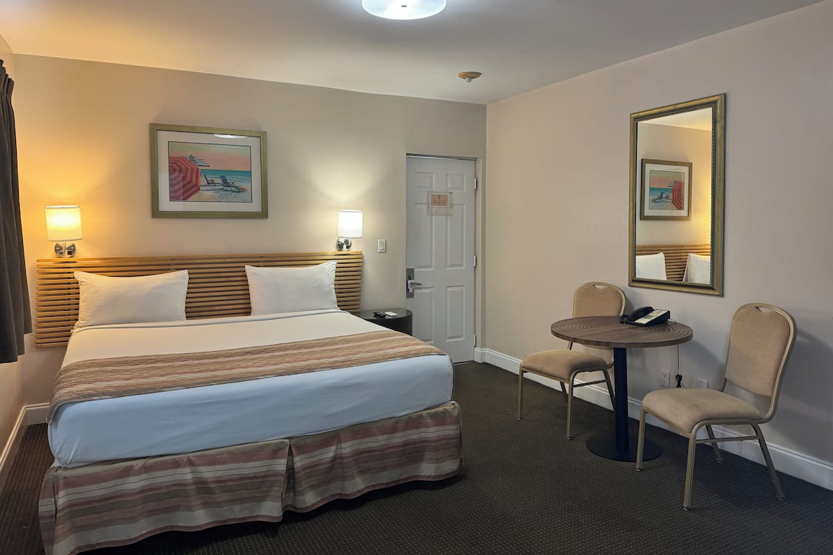 A hotel room with a bed, bedside lamps, a round table with a chair, a mirror, and a door.
