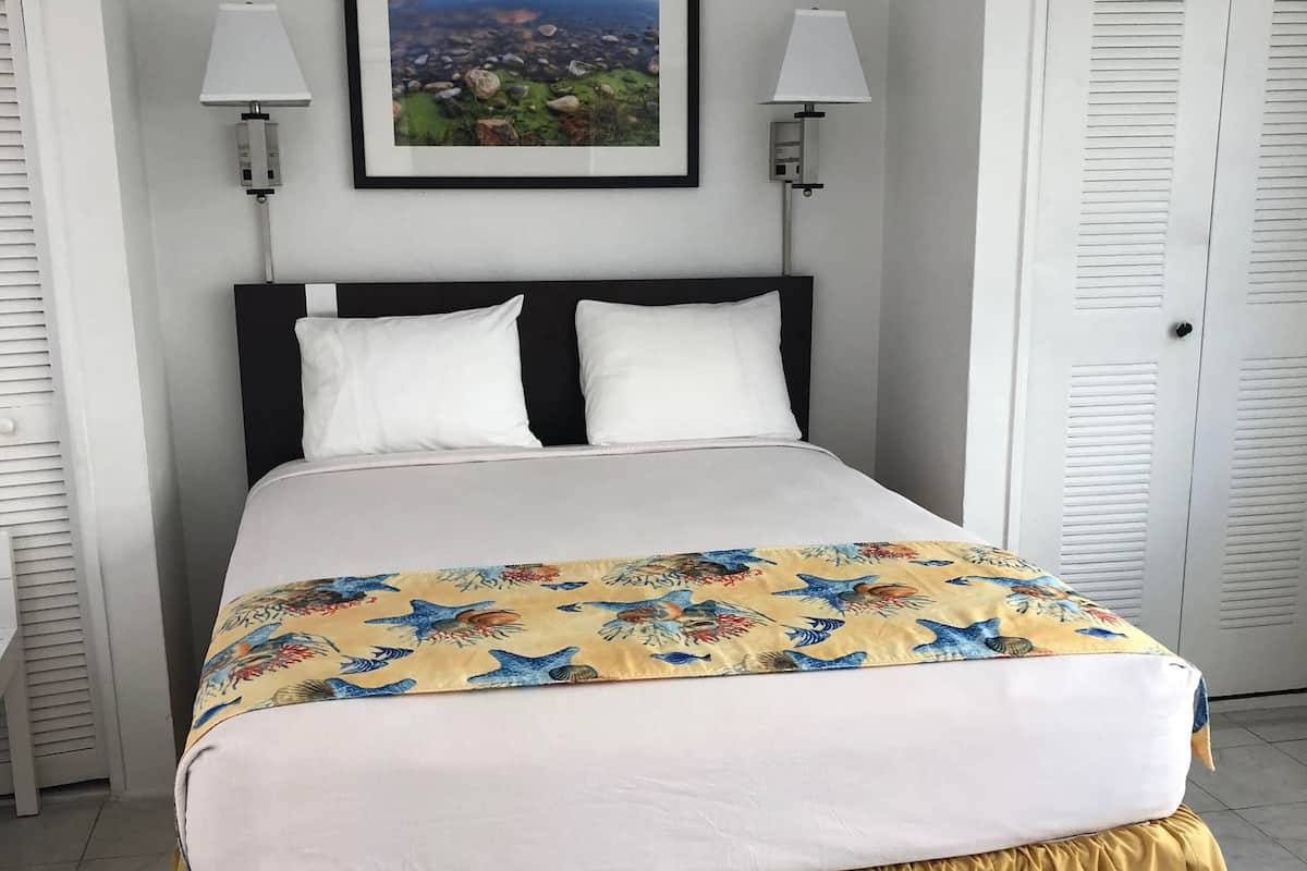 A neatly made bed with a floral bedspread and a yellow skirted duvet. There are two wall-mounted lamps on either side of the bed, and a framed landscape painting hangs above it.