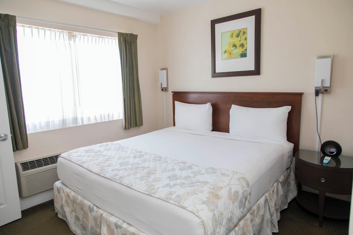 A hotel room with a large bed, a bedside table with a clock, a wall-mounted air conditioner, and a framed picture on the wall.
