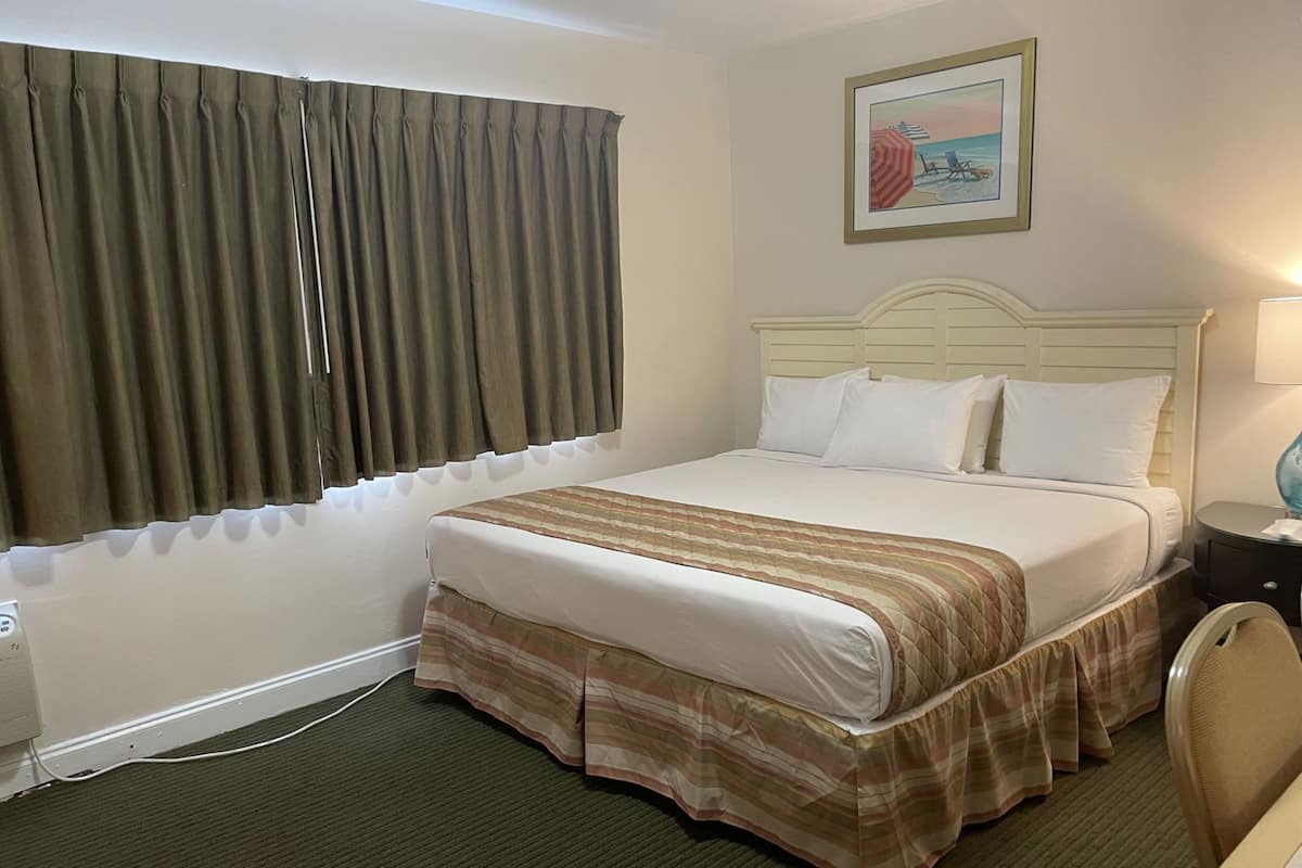 A hotel room with a bed, a nightstand, a lamp, a window with curtains, and a painting on the wall.