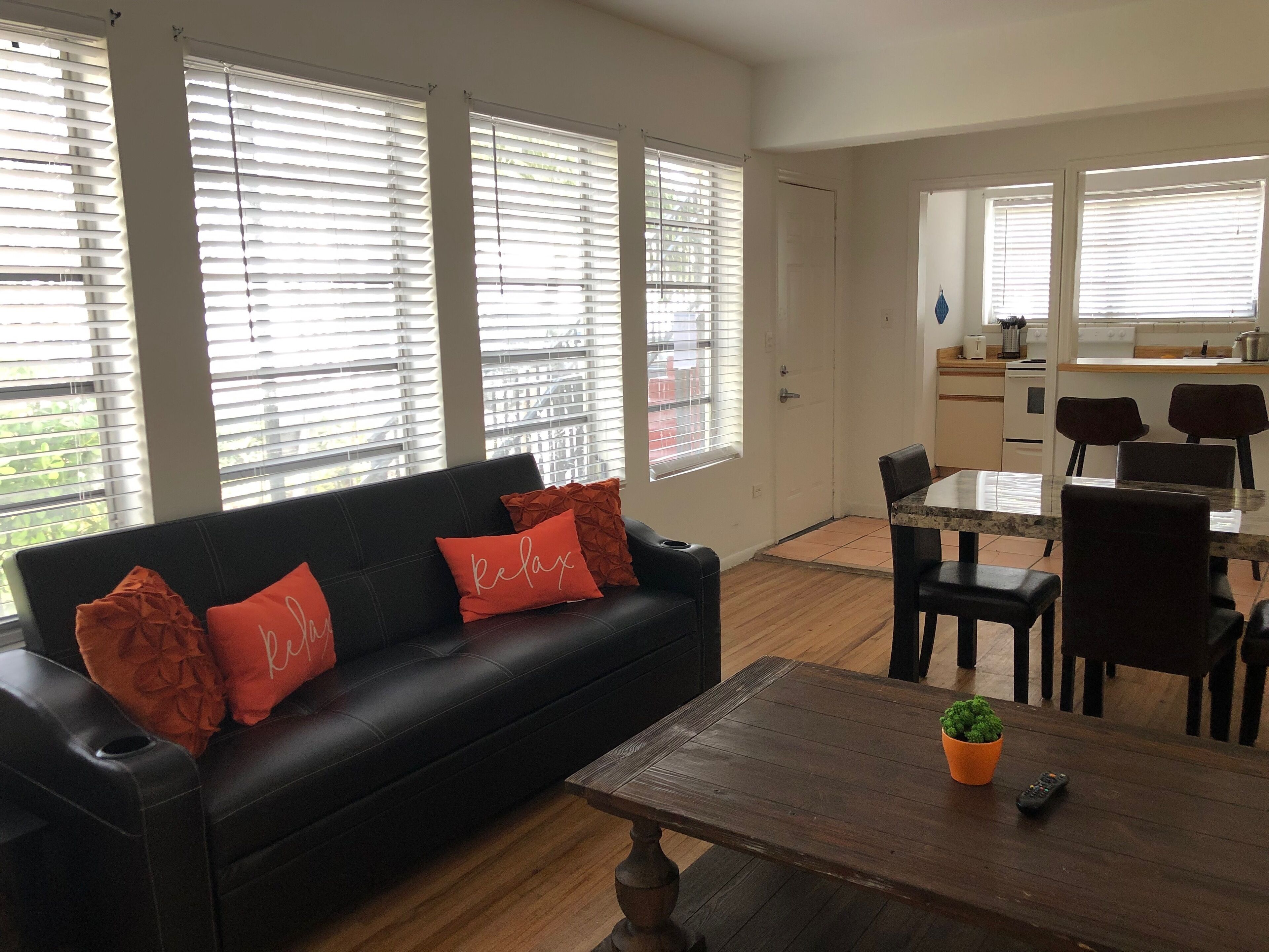A modern living room with a black leather sofa, orange cushions, a wooden coffee table, and a dining area with a table and chairs.