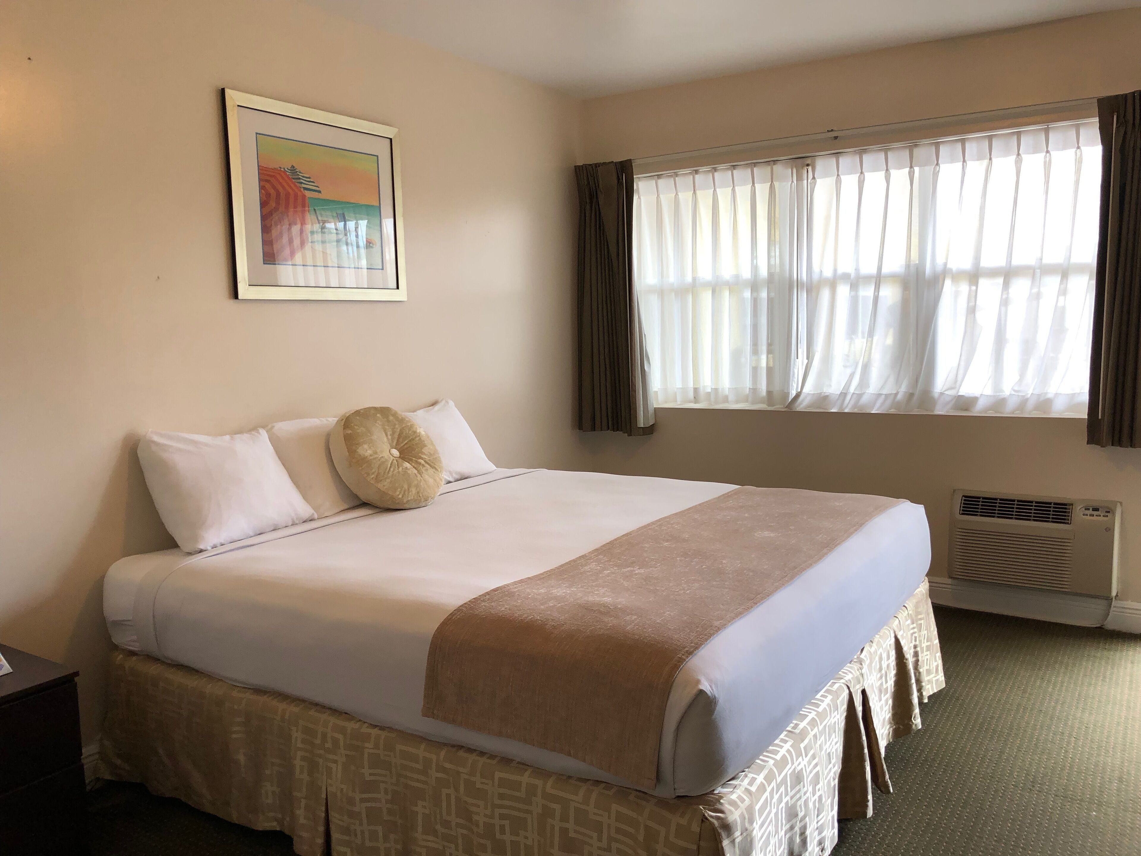 A hotel room with a bed, a window with curtains, a wall-mounted air conditioner, and a framed picture.