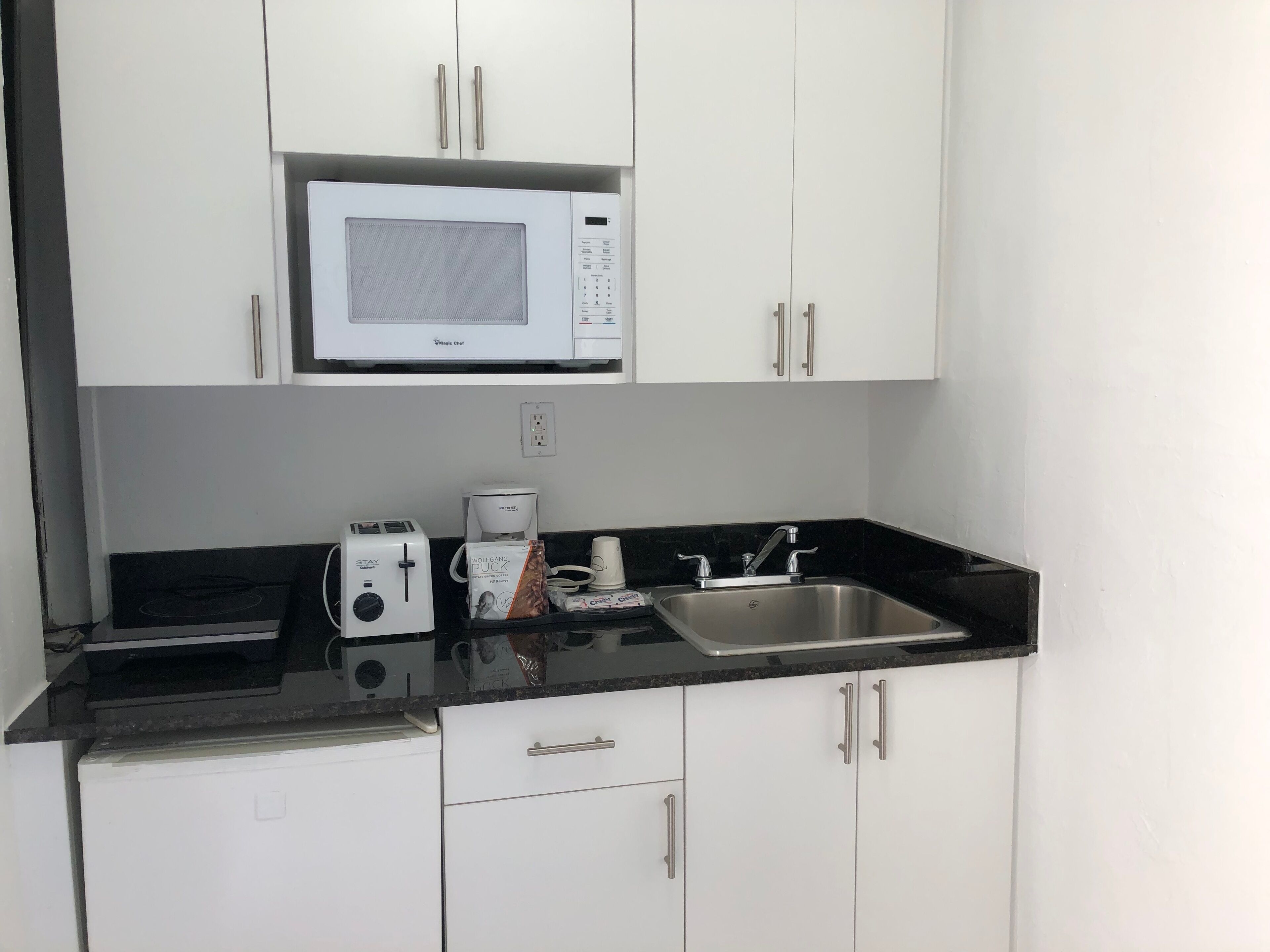 A compact kitchen with white cabinets, a microwave, a toaster, a coffee maker, and a sink.