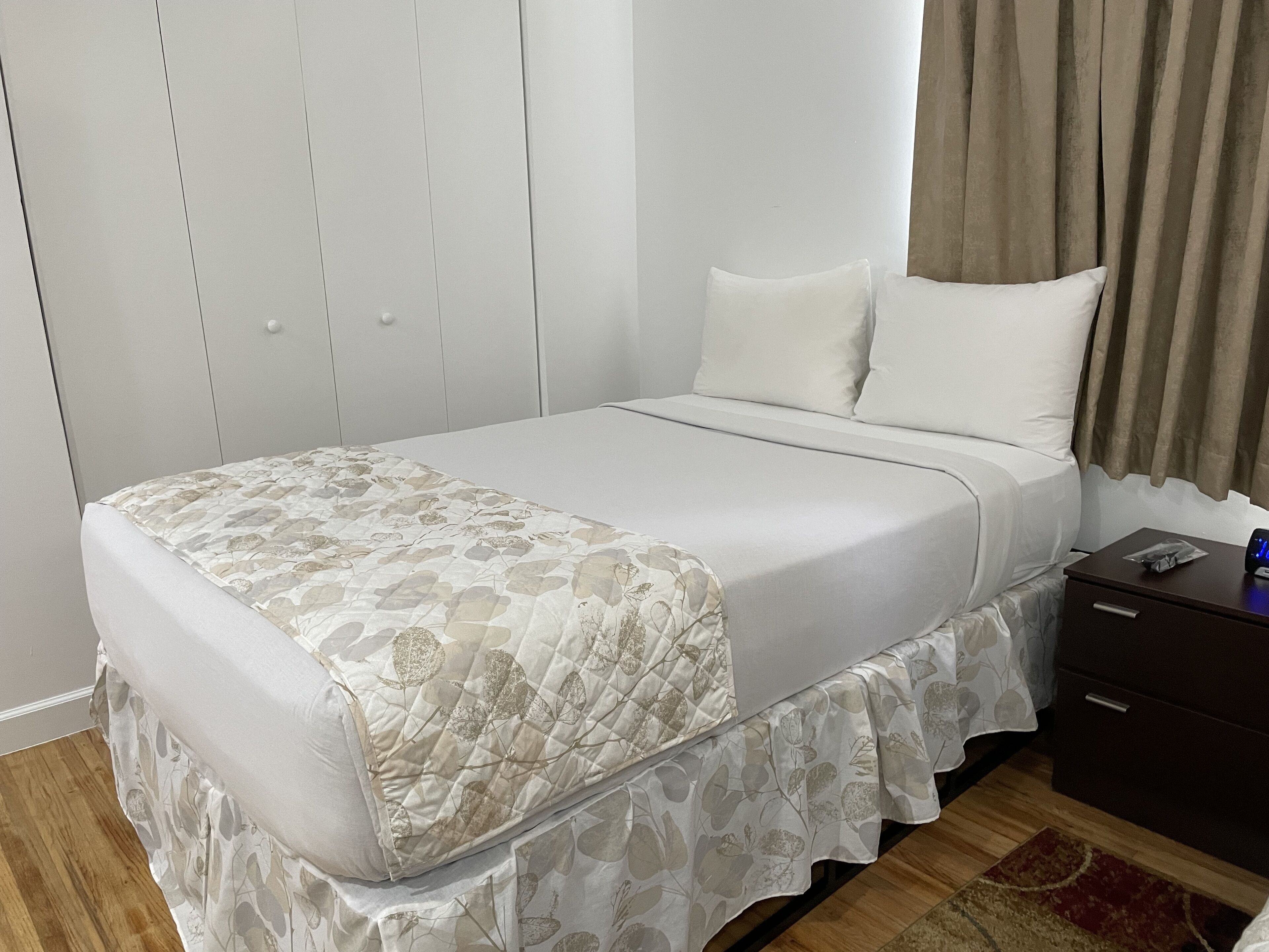 A neatly made bed with a floral bedspread, a bedside table with a phone and a small item on it, and a wooden floor.
