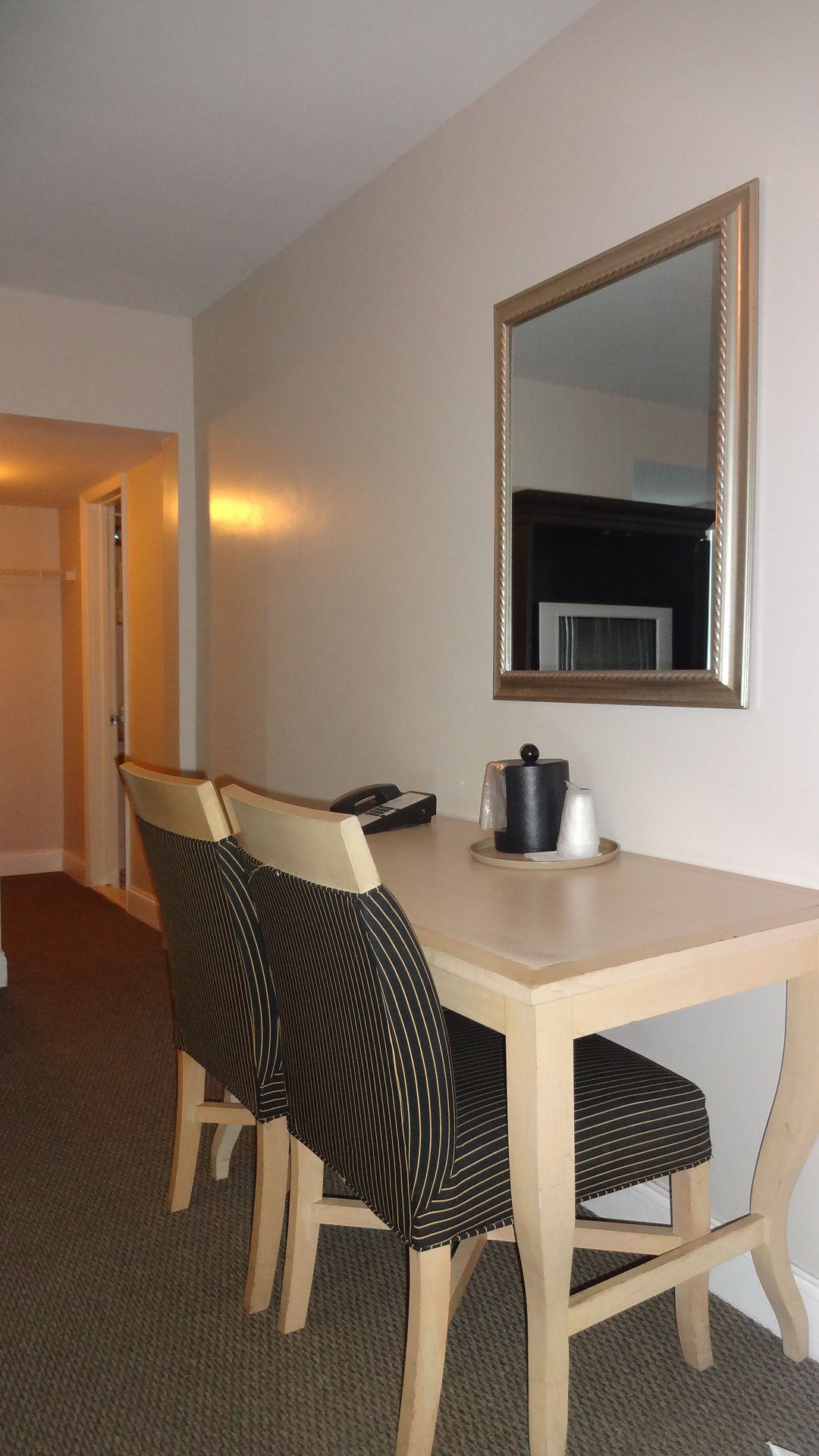 Habitación de hotel con una mesa de comedor de madera, dos sillas a rayas, una cafetera y un espejo en la pared.