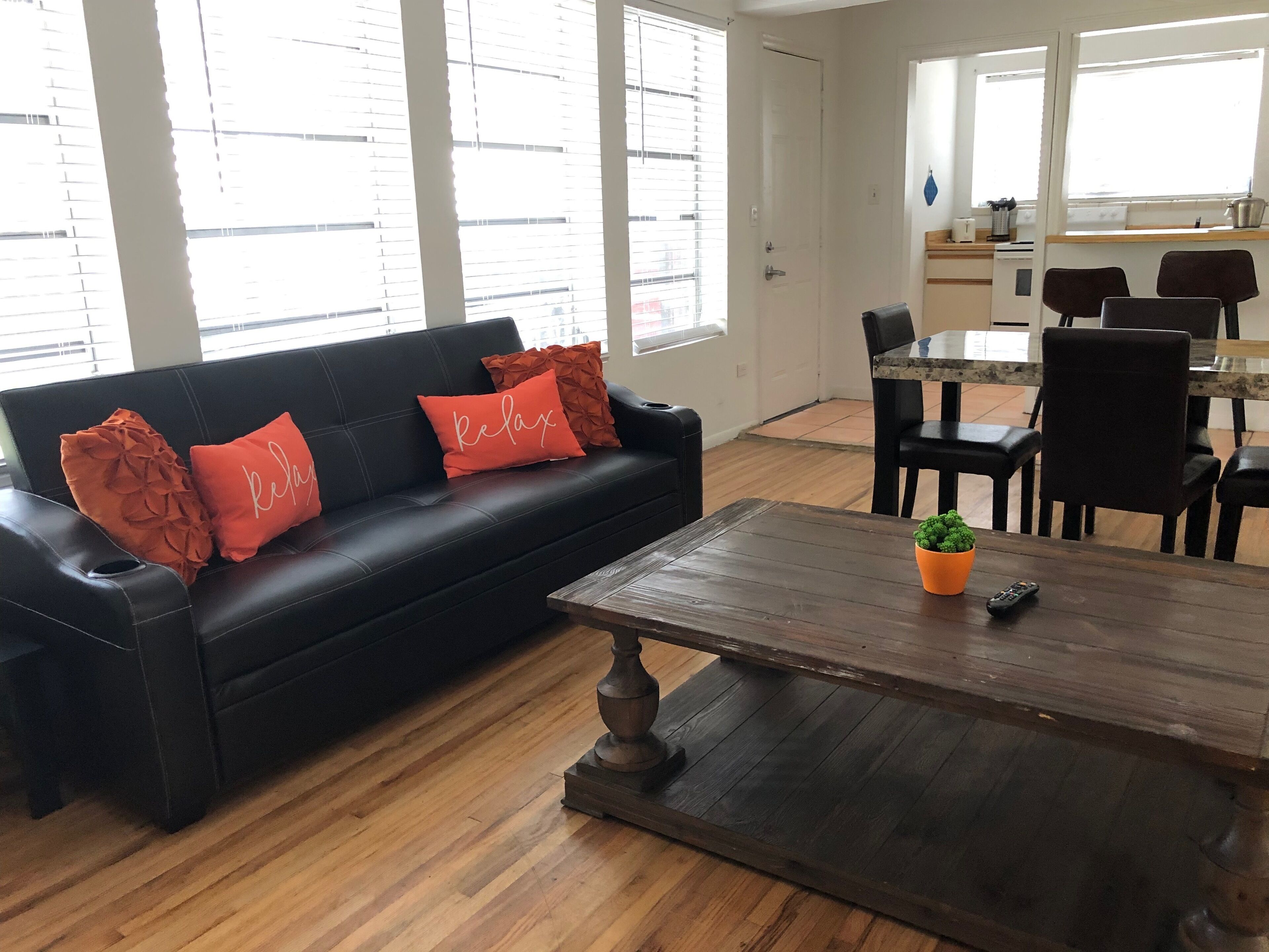 A modern living room with a black leather sofa, two orange pillows, a wooden coffee table, and a small potted plant.