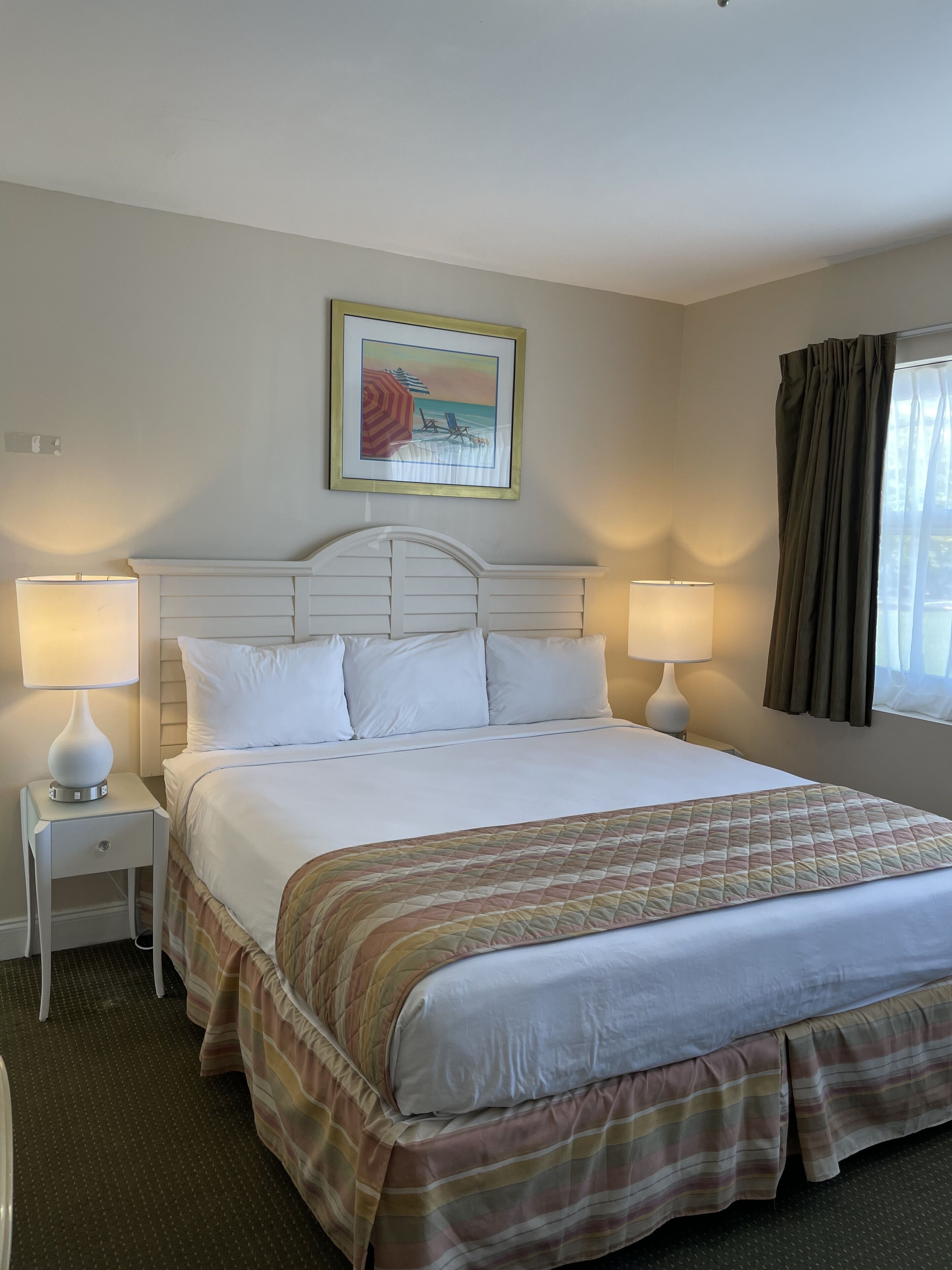 A hotel room with a large bed, two bedside lamps, a painting of a beach scene, and a window with curtains.