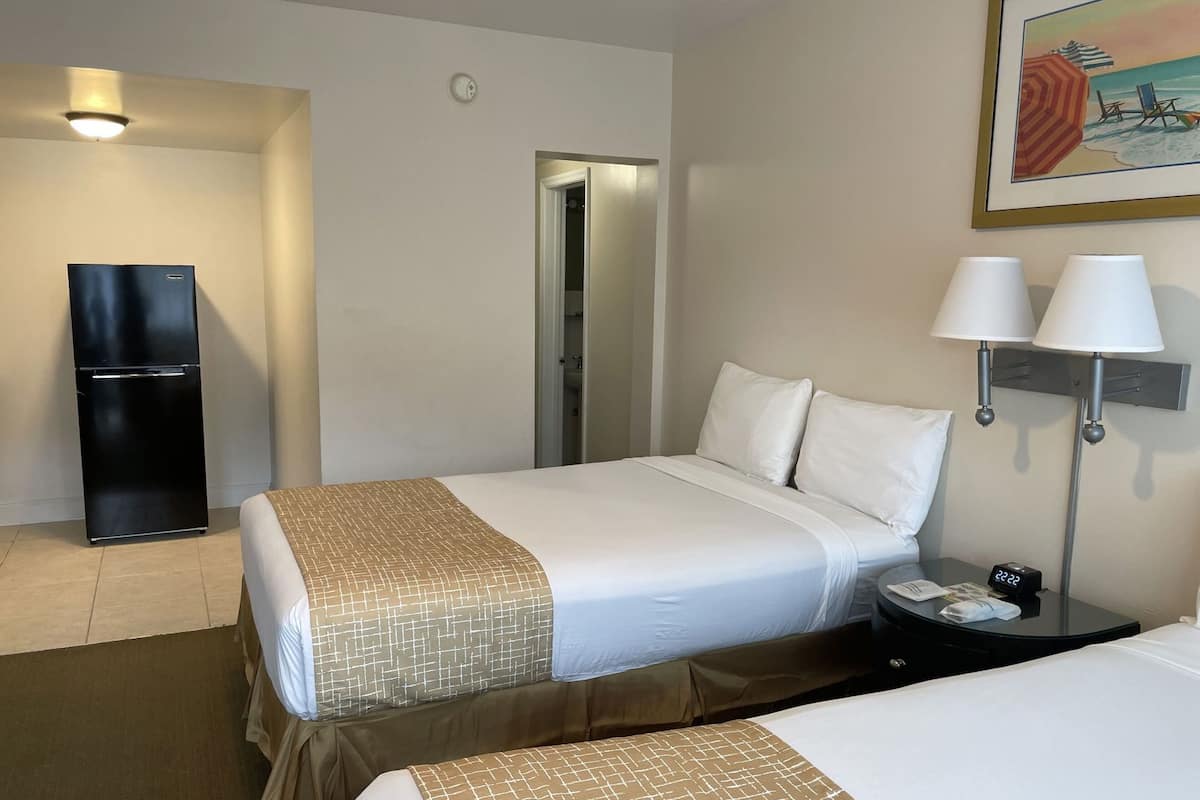 A hotel room with a bed, a bedside table with a lamp, a refrigerator, and a painting on the wall.