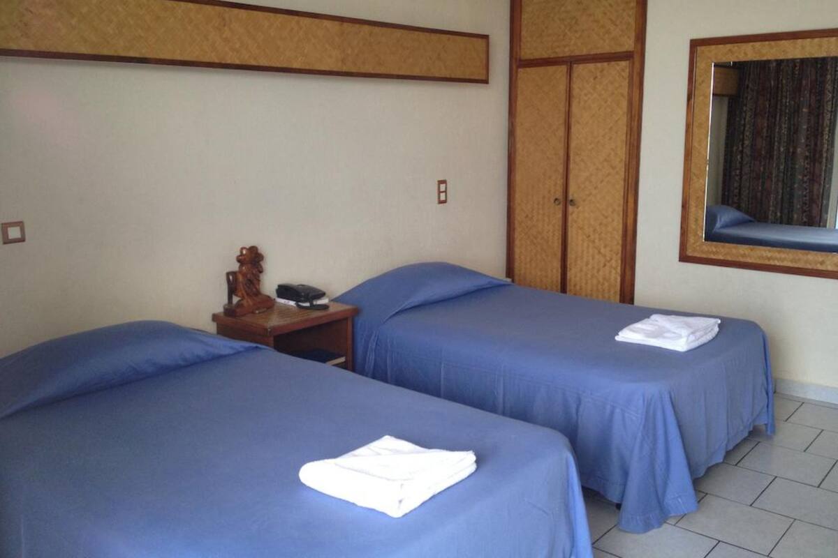 A hotel room with two single beds, a wooden nightstand with a telephone and a decorative item, a wardrobe, and a window with a view.