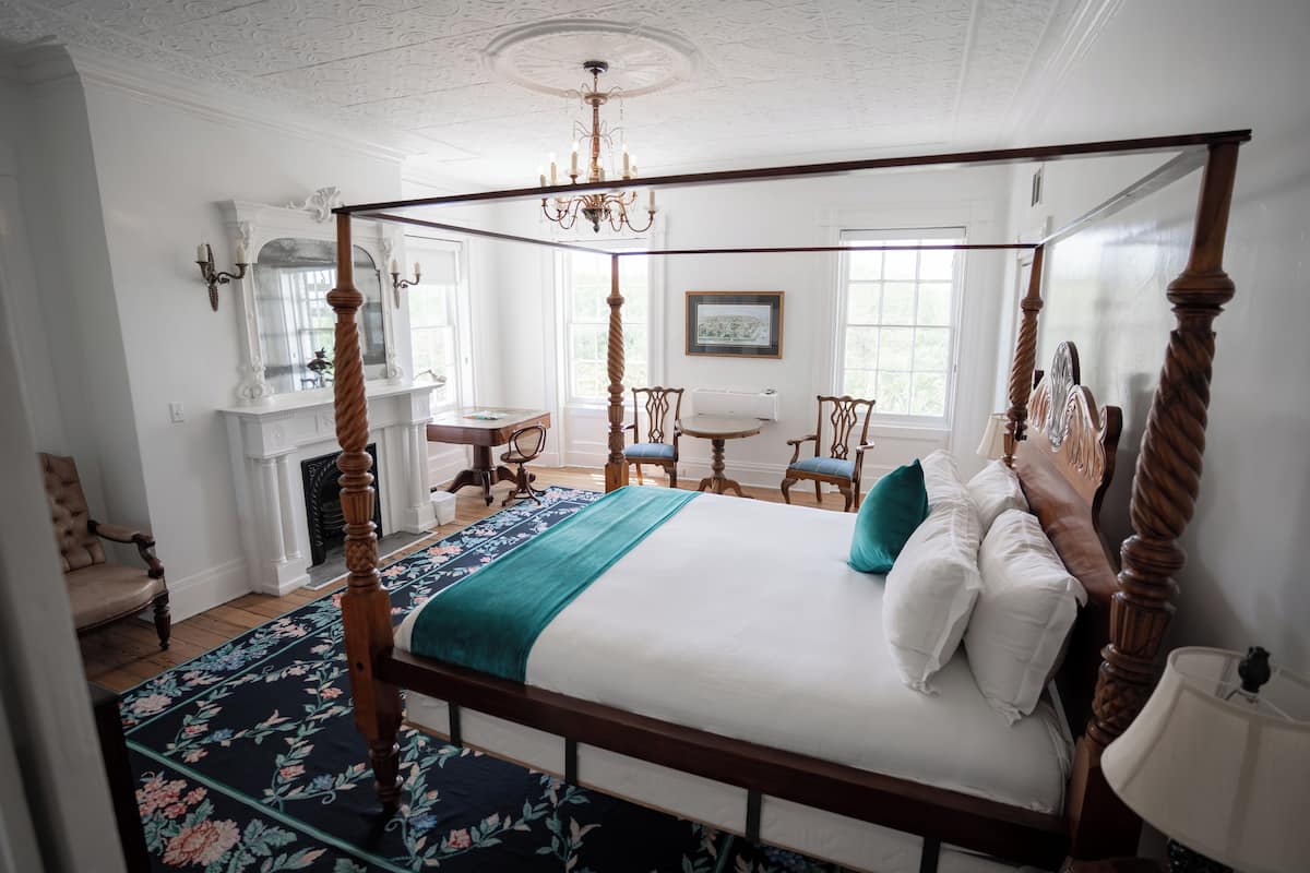 A four-poster bed with a floral bedspread, a chandelier, and a view of the outdoors through large windows.