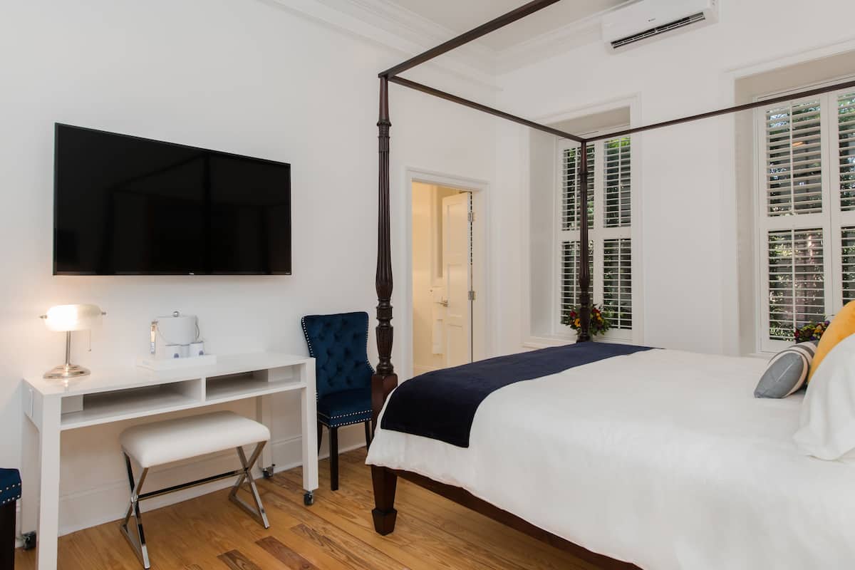 A bedroom with a four-poster bed, a desk with a chair, a flat-screen TV, and a window with shutters.