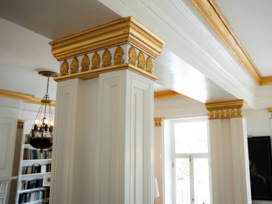Et værelse med en hvid søjle med guld detaljer, en lysekrone og en reol i baggrunden.