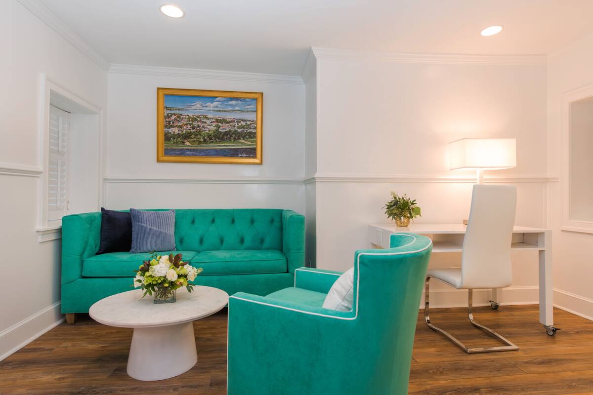 A room with a teal sofa, a white armchair, a small round table with a vase of flowers, a desk with a lamp, and a framed landscape painting on the wall.