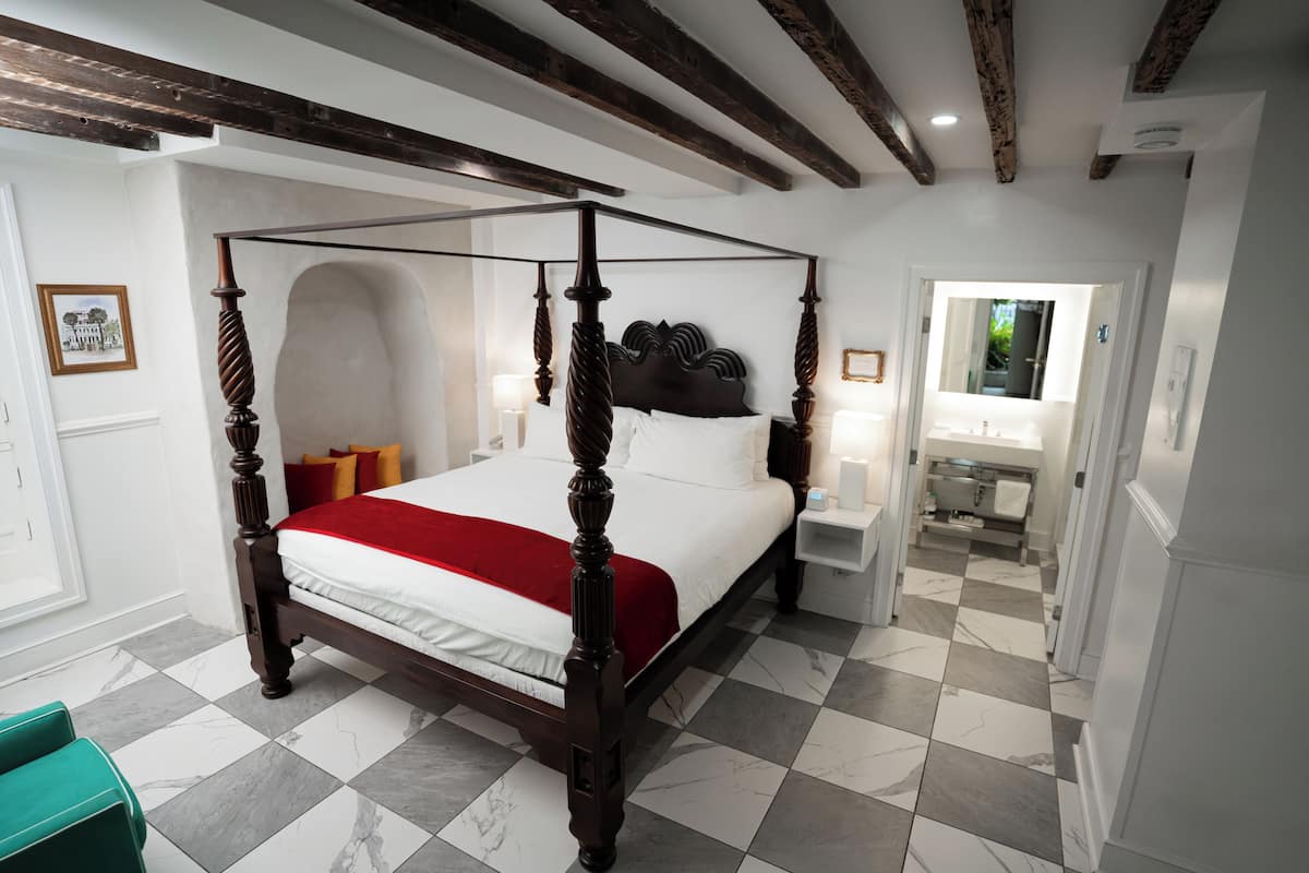 A four-poster bed with a red throw, white linens, and a checkered floor. There is a nightstand with a lamp, a mirror, and a view into a bathroom with a sink and mirror.