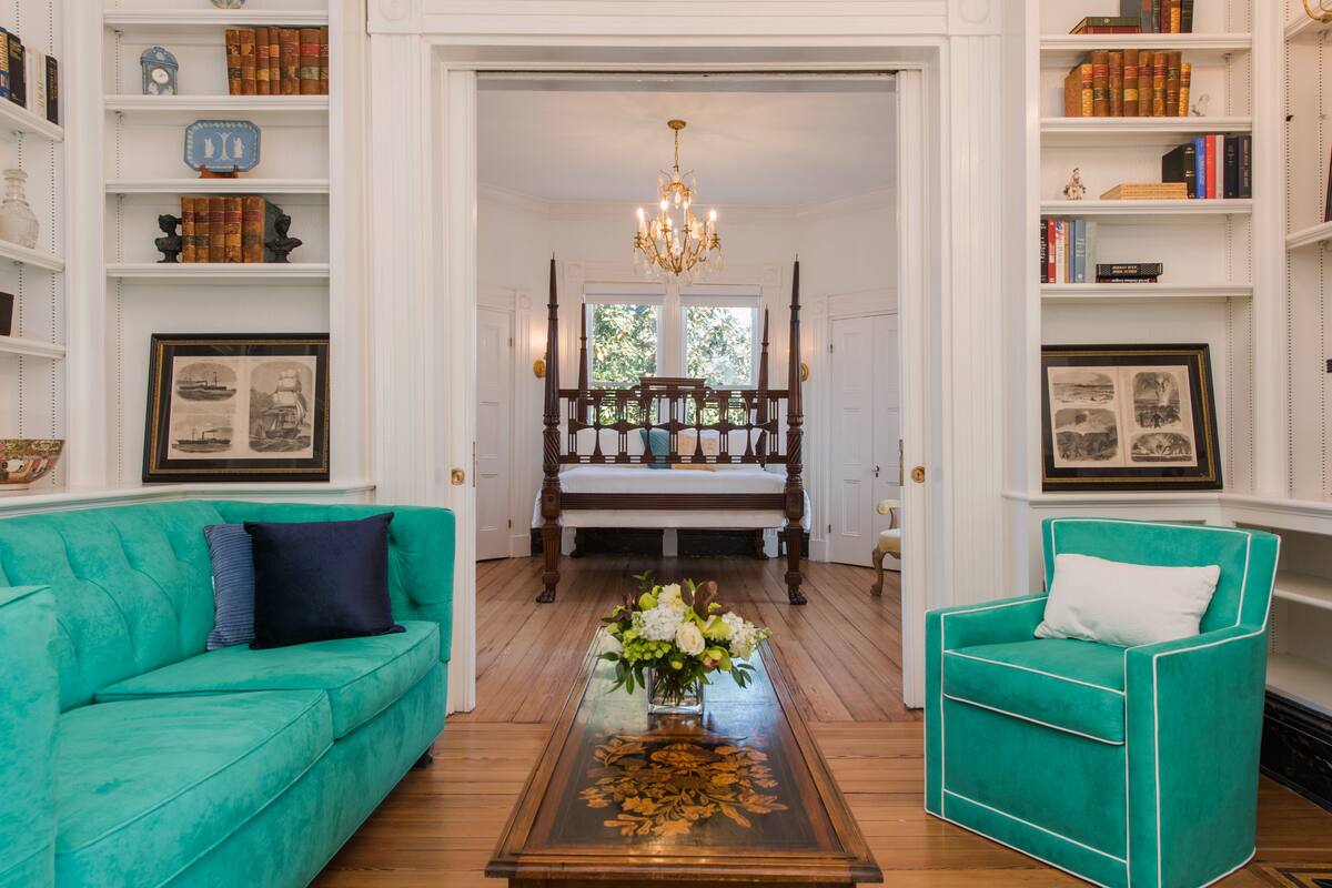 A room with a teal sofa, a matching armchair, a wooden coffee table with a floral centerpiece, and a large window with a view of trees.