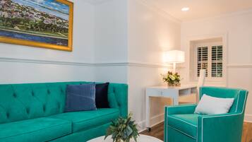 A teal-colored sofa and armchair in a room with white walls, a round white table, and a painting of a coastal town.