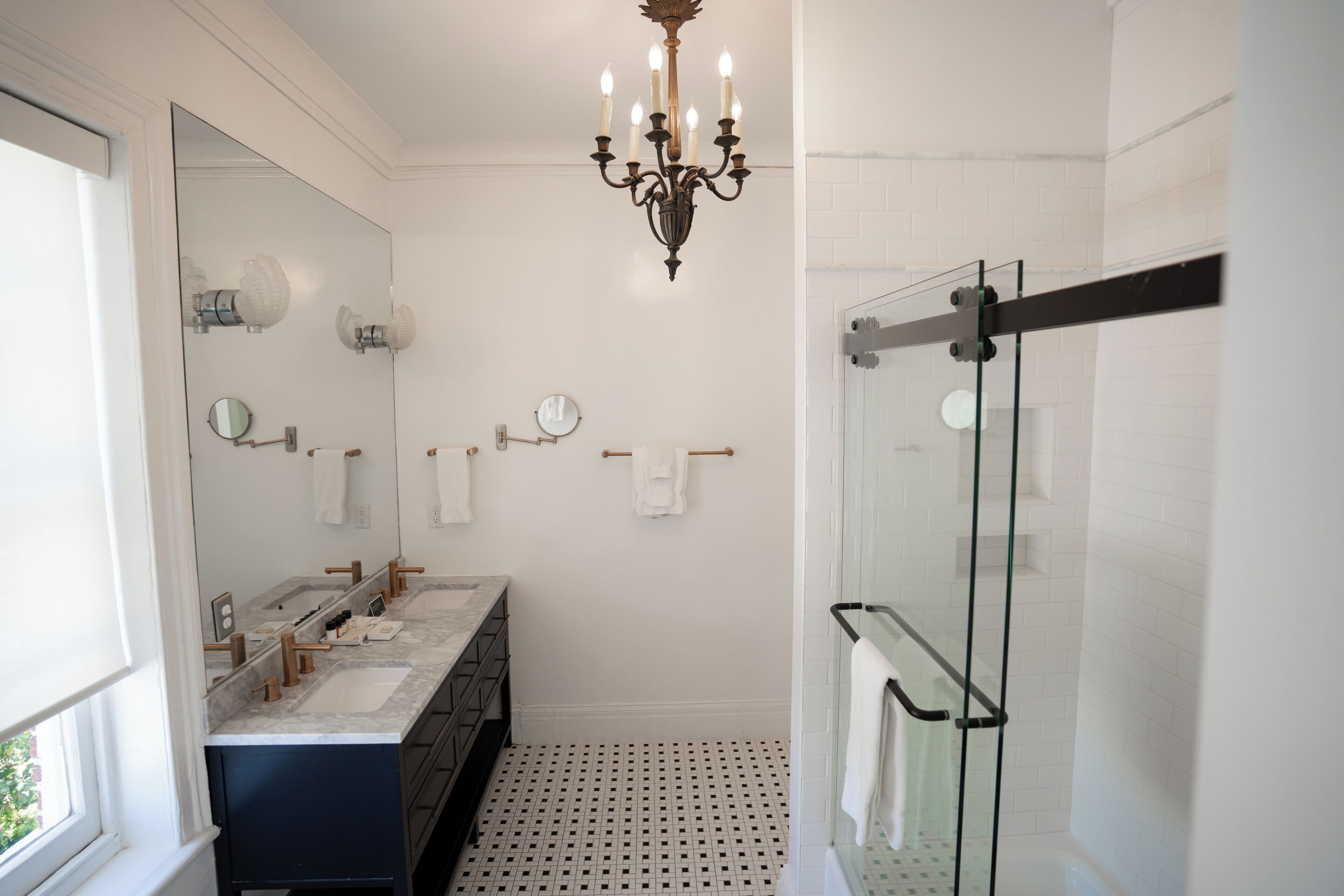 A modern bathroom with a large mirror, two sinks, a chandelier, and a glass-enclosed shower.