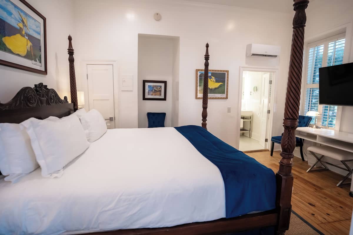 A four-poster bed with white and blue bedding, a wooden desk with a chair, a television, and framed artwork on the walls.