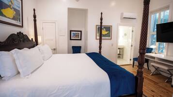 A four-poster bed with white and blue bedding, a wooden desk with a chair, a television, and framed artwork on the walls.