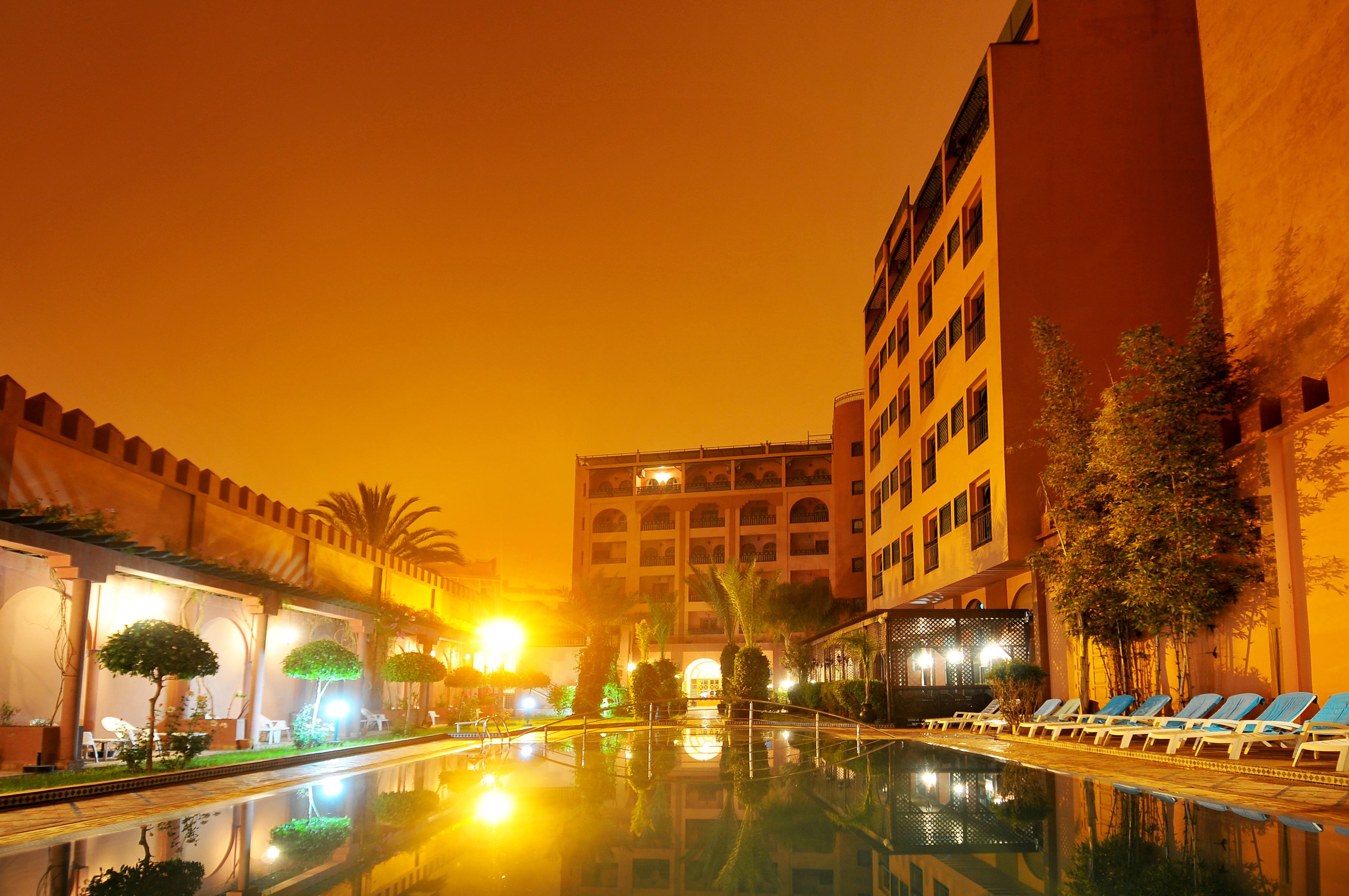 Zona de piscina de hotel con sillas de descanso y un estanque reflectante, rodeada de palmeras y edificios.