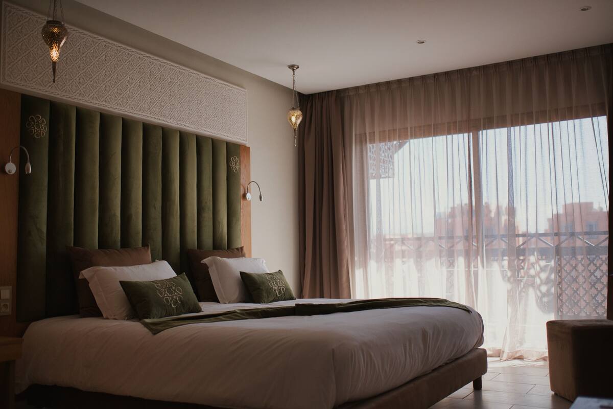 Moderni hotellihuone, jossa on suuri sänky, pystysuorilla paneeleilla varustettu pääty, suuri ikkuna läpikuultavilla verhoilla ja parveke, jolta on näkymä.