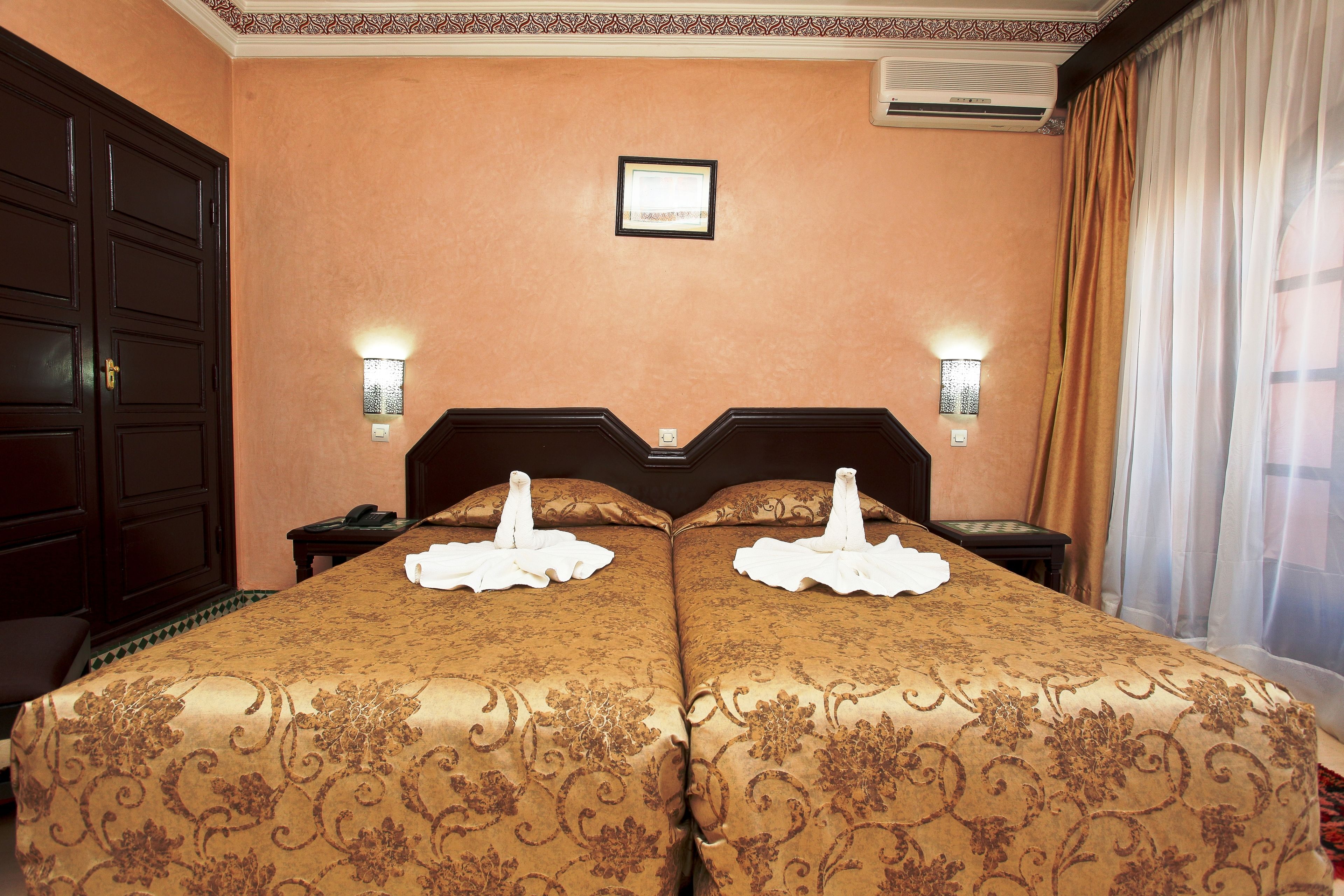 Un lit double avec une couvre-lit à motifs floraux, deux tables de chevet avec des lampes, un tableau encadré au mur et une grande fenêtre avec des rideaux.