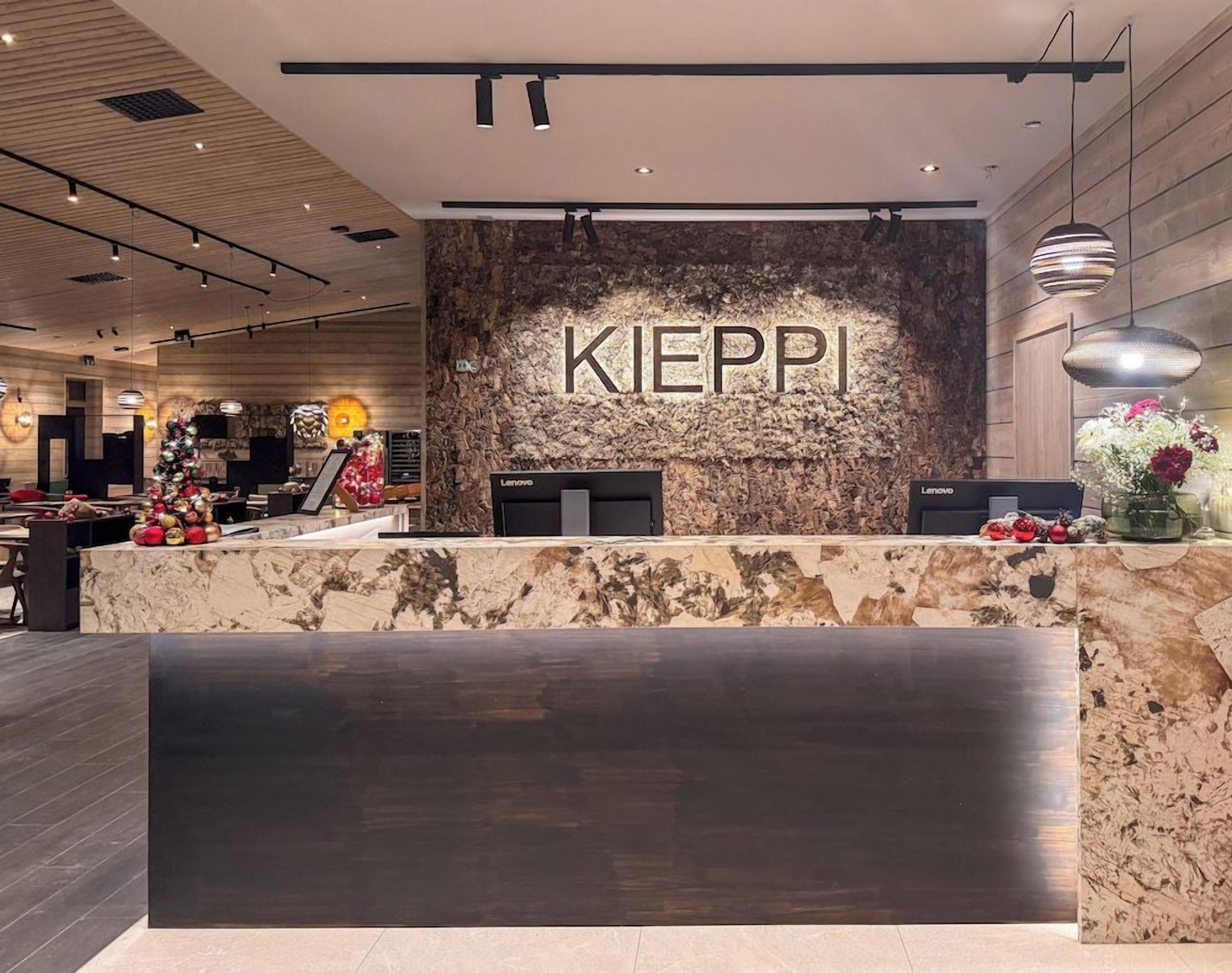 A reception desk with the word 'KIEPI' on a textured wall, a modern chandelier, and a counter with a vase of flowers.