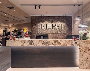 A reception desk with the word 'KIEPI' on a textured wall, a modern chandelier, and a counter with a vase of flowers. - Hotelli Kieppi (Saariselka)