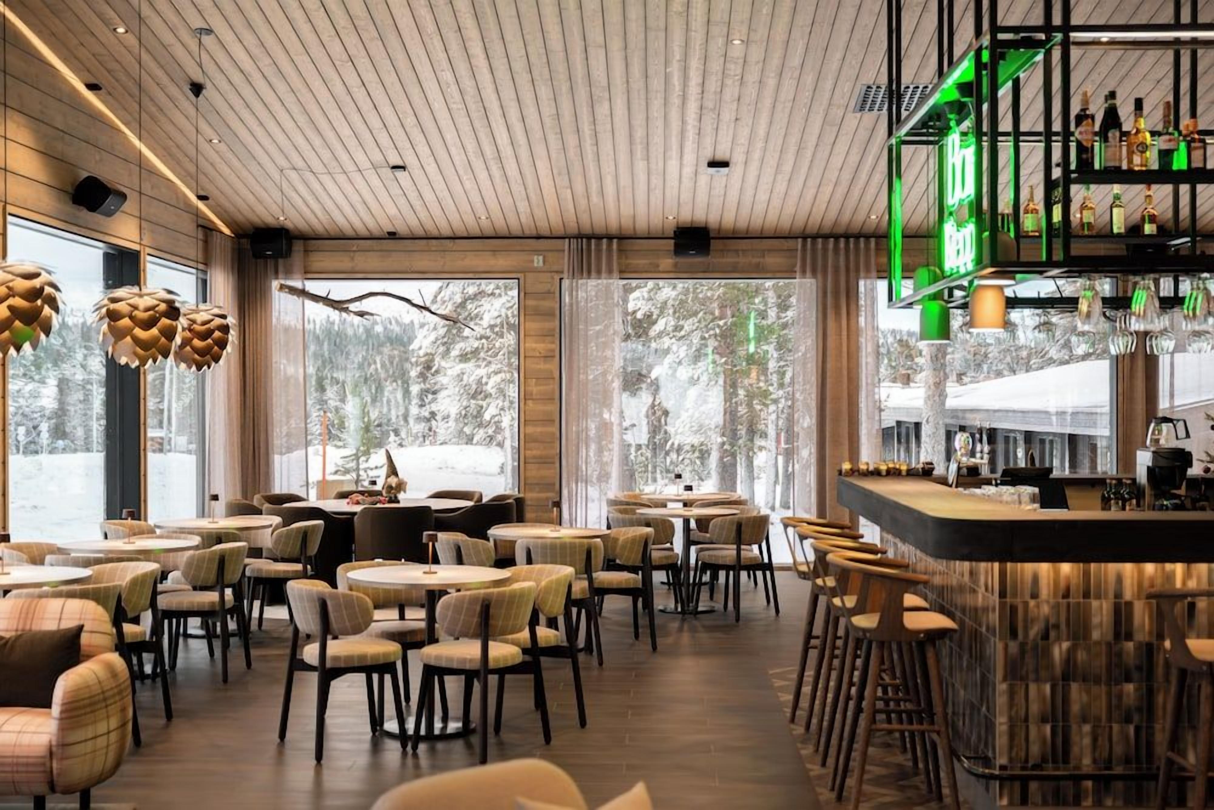 A restaurant with a bar area, wooden ceiling, and large windows with a view of trees.