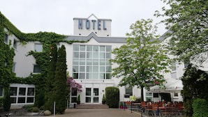Un hotel con un amplio ventanal de vidrio, un letrero que dice "HOTEL" sobre la entrada, y área de estar exterior con mesas y sillas.