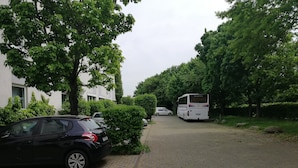 Un camino pavimentado con coches aparcados y un autobús, rodeado de árboles y un edificio de fachada blanca.