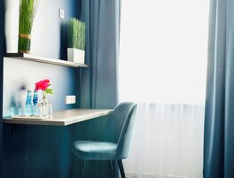 Una habitación moderna con una silla azul, una mesita con objetos decorativos y vistas por una ventana con cortinas.