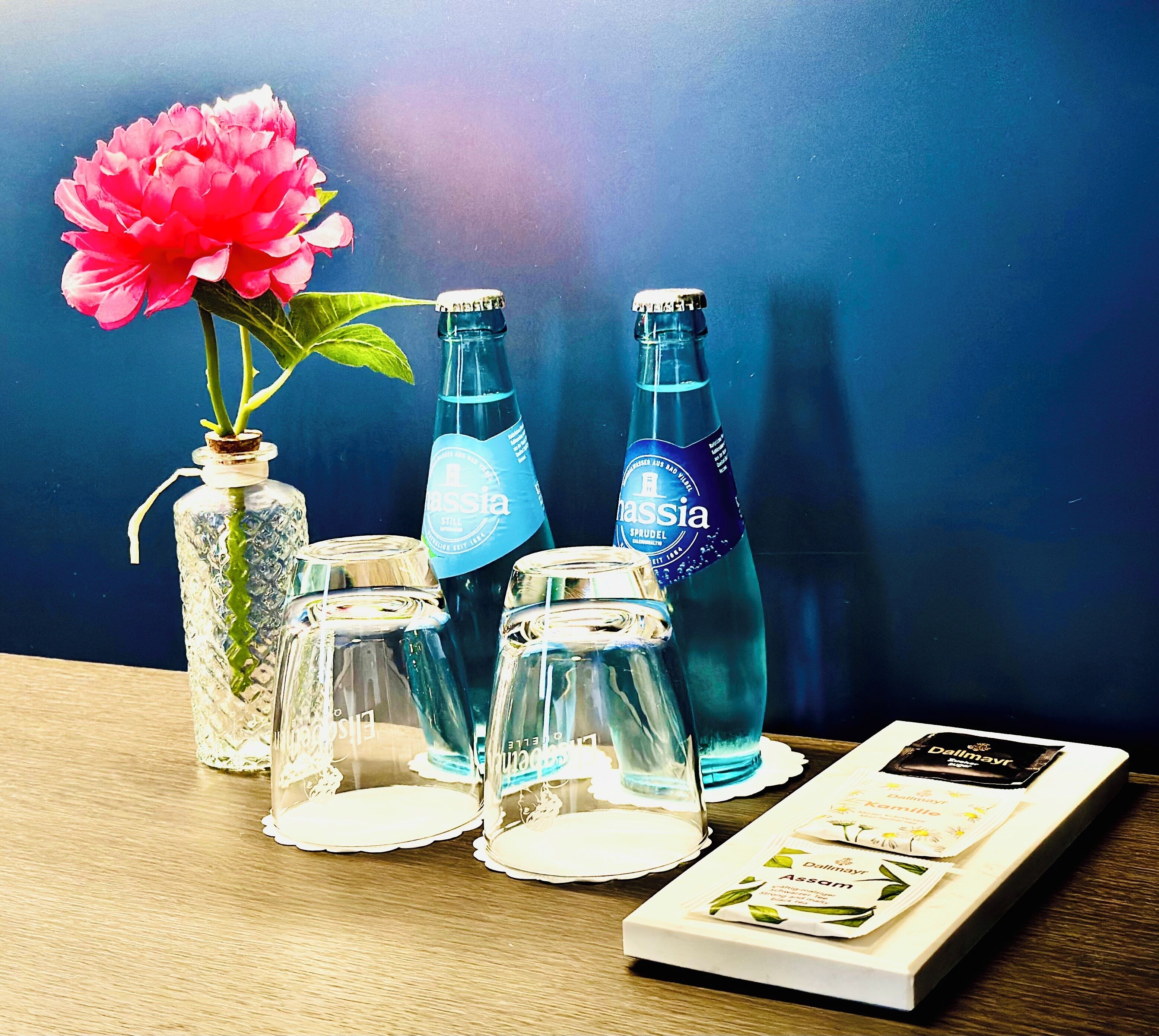 Una mesa con un florero de vidrio con una flor rosa, dos botellas de agua y un folleto sobre una superficie de madera, sobre un fondo azul.