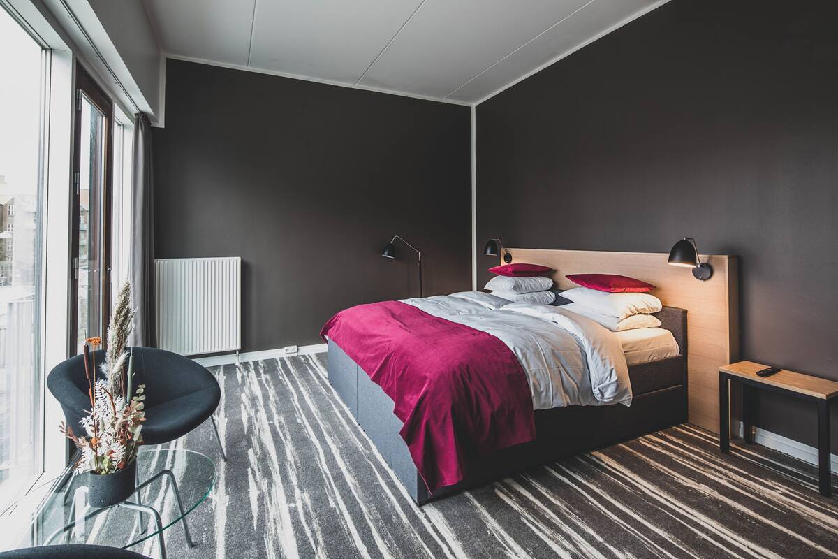 A modern bedroom with a large bed, a side table, a chair, and a view of the city through large windows.