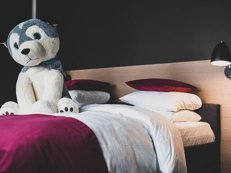 Una cama con un peluche, almohadas y una lámpara de noche, además de un cabecero.
